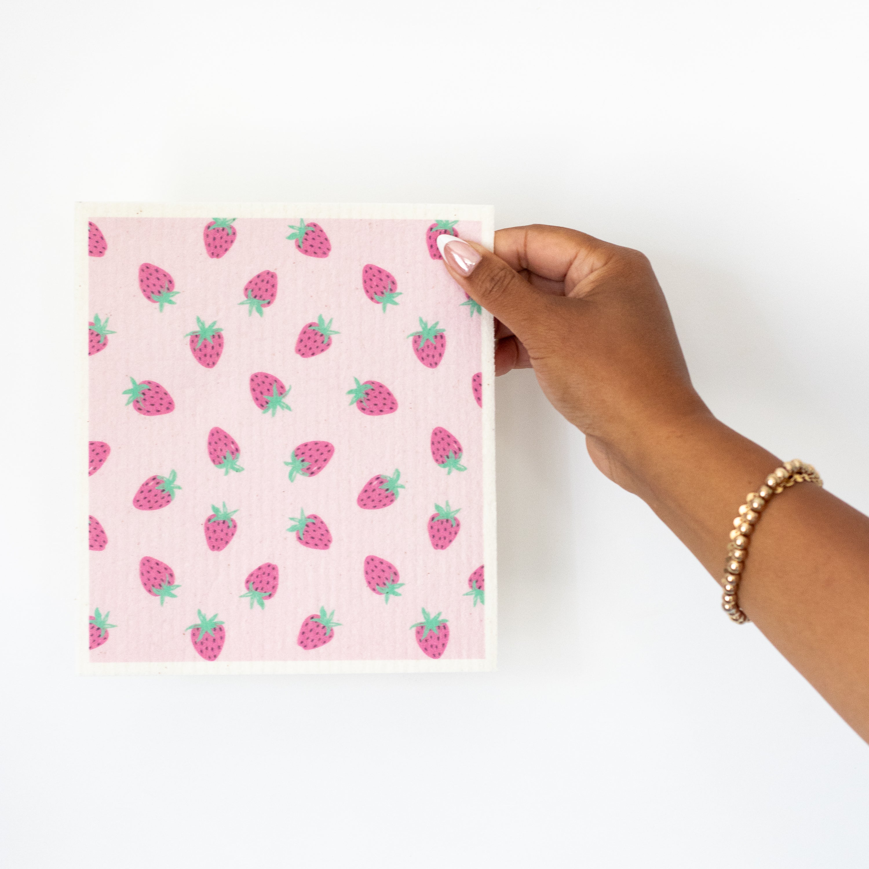 A hand holding a Swedish dishcloth featuring a pink strawberry pattern against a plain white background.