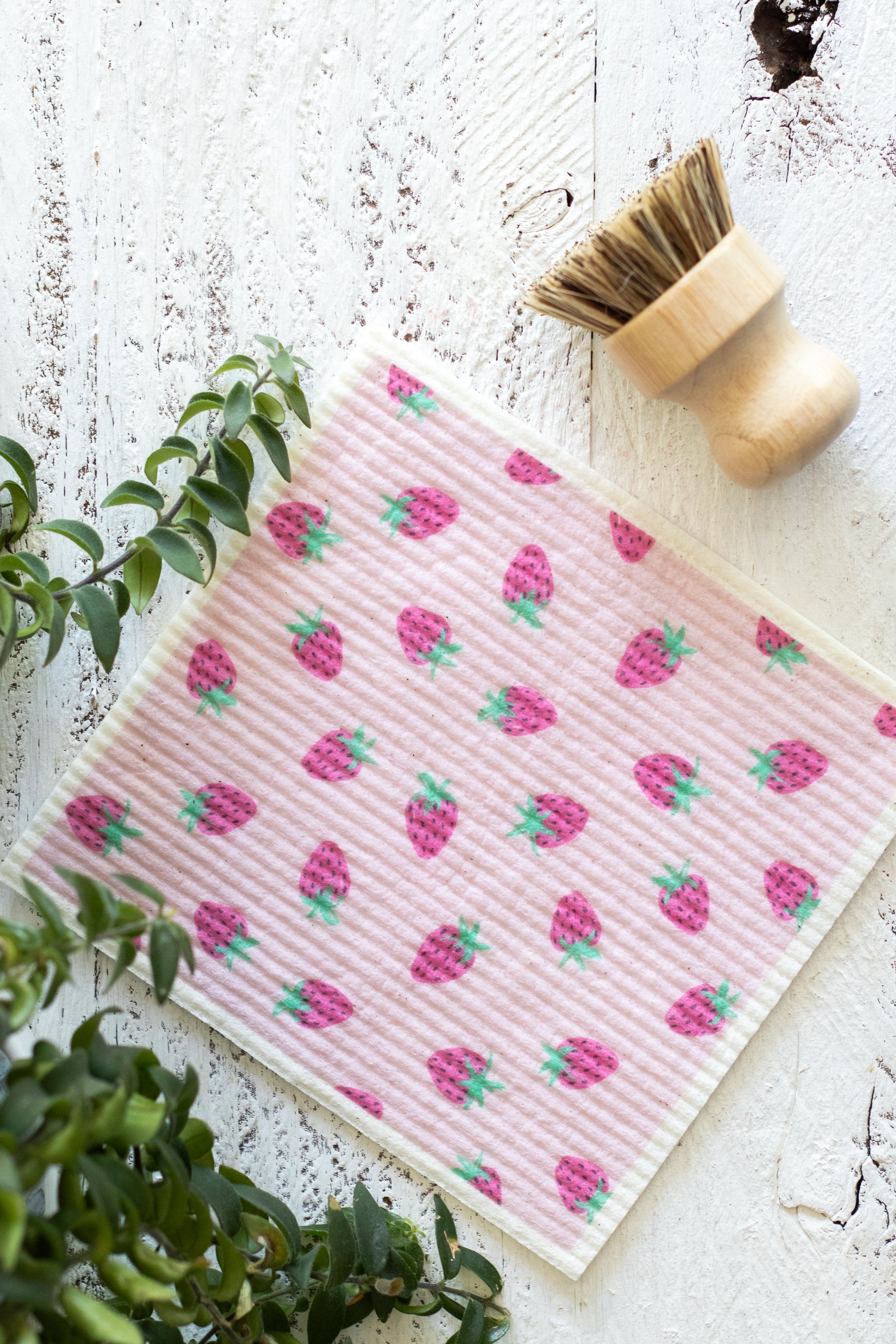 A flat-lay view of a pink Swedish dishcloth with a repeating strawberry pattern, styled on a distressed white wooden surface alongside a wooden-handled cleaning brush and green foliage.