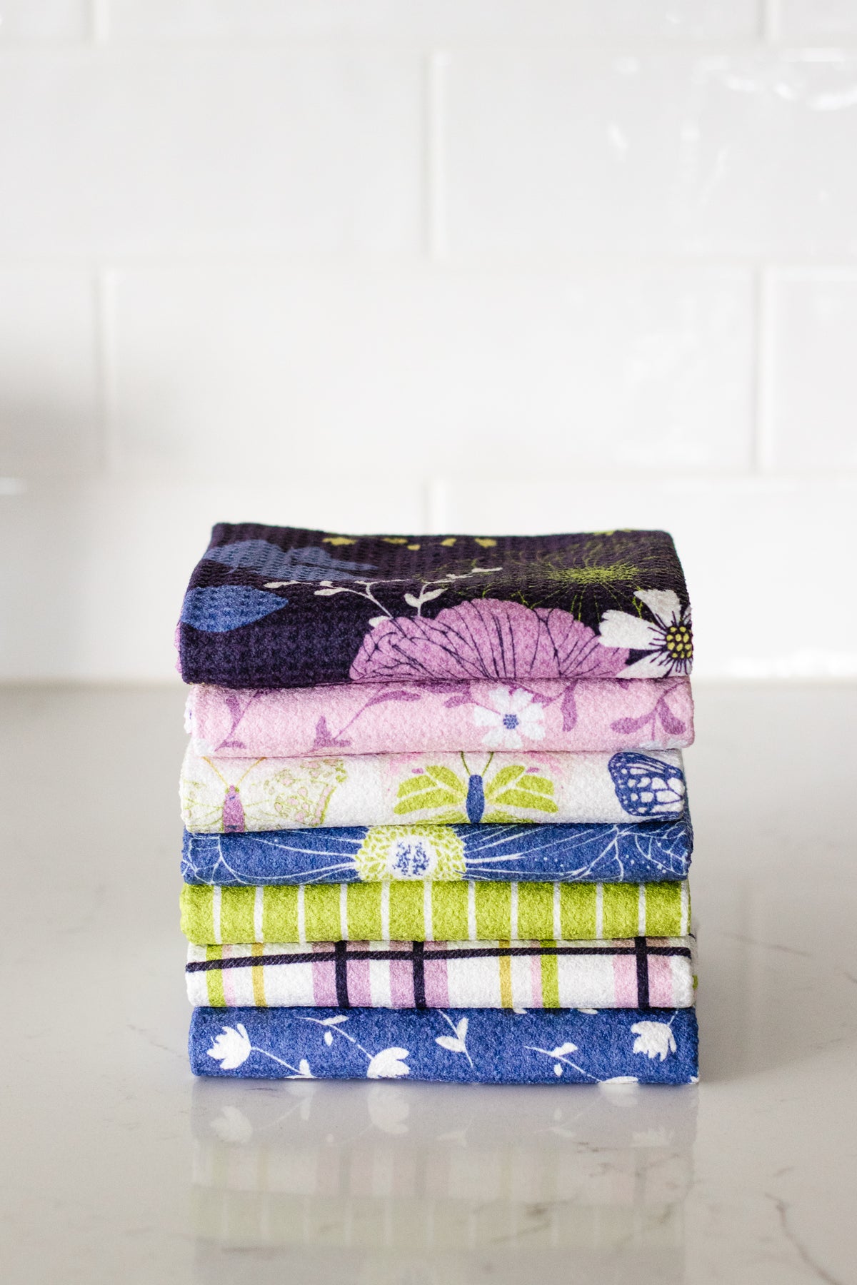 A stack of seven folded kitchen tea towels with various colorful patterns, including floral, stripes, and plaid prints, displayed on a light marble surface against a white subway tile background.