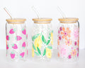 Three can-style glass tumblers that have colorful floral and fruit designs, with bamboo lids and glass straws, on a white background.