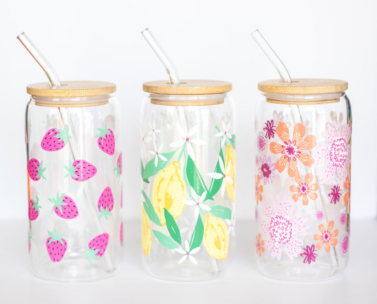 Three can-style glass tumblers that have colorful floral and fruit designs, with bamboo lids and glass straws, on a white background.