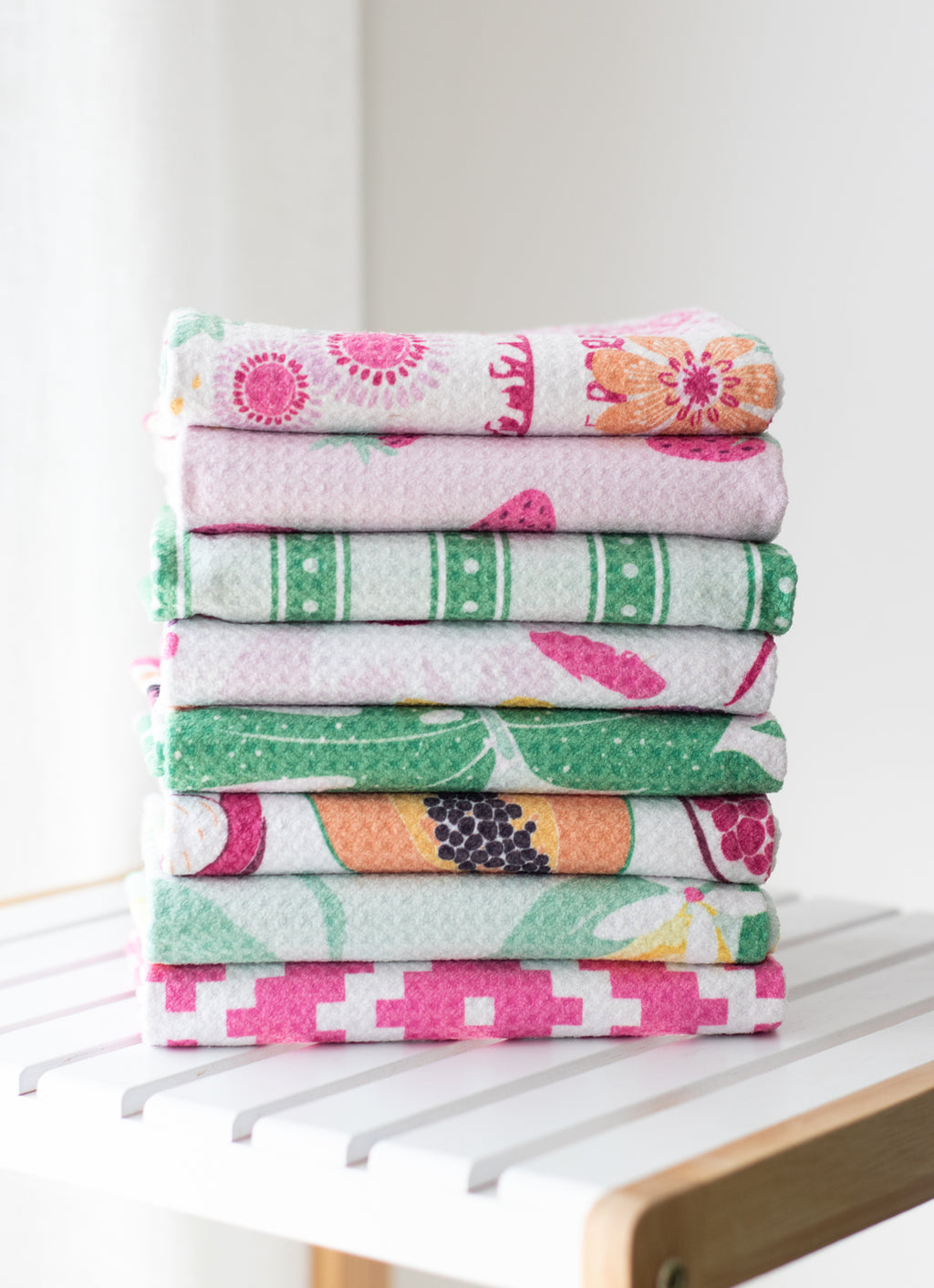 A stack of eight folded waffle-weave kitchen towels with various colorful floral and geometric patterns, displayed on a white slatted wooden table against a bright, neutral background.