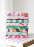 A stack of eight folded waffle-weave kitchen towels with various colorful floral and geometric patterns, displayed on a white slatted wooden table against a bright, neutral background.