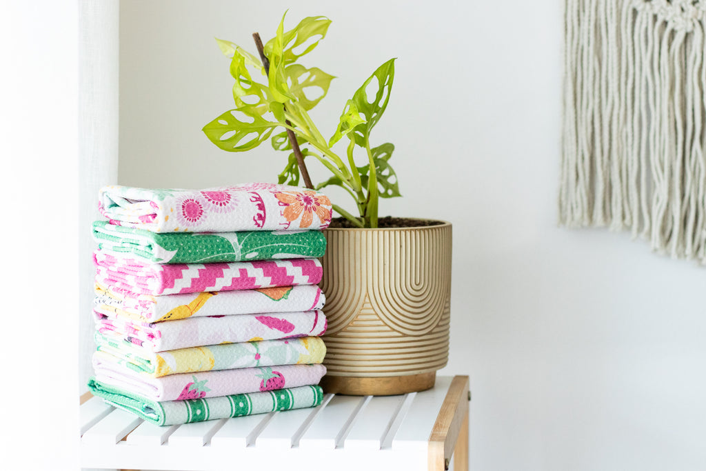 A stack of folded waffle-weave kitchen tea towels sits on a white slatted side table next to a potted green plant in a ribbed beige pot, against a white wall.