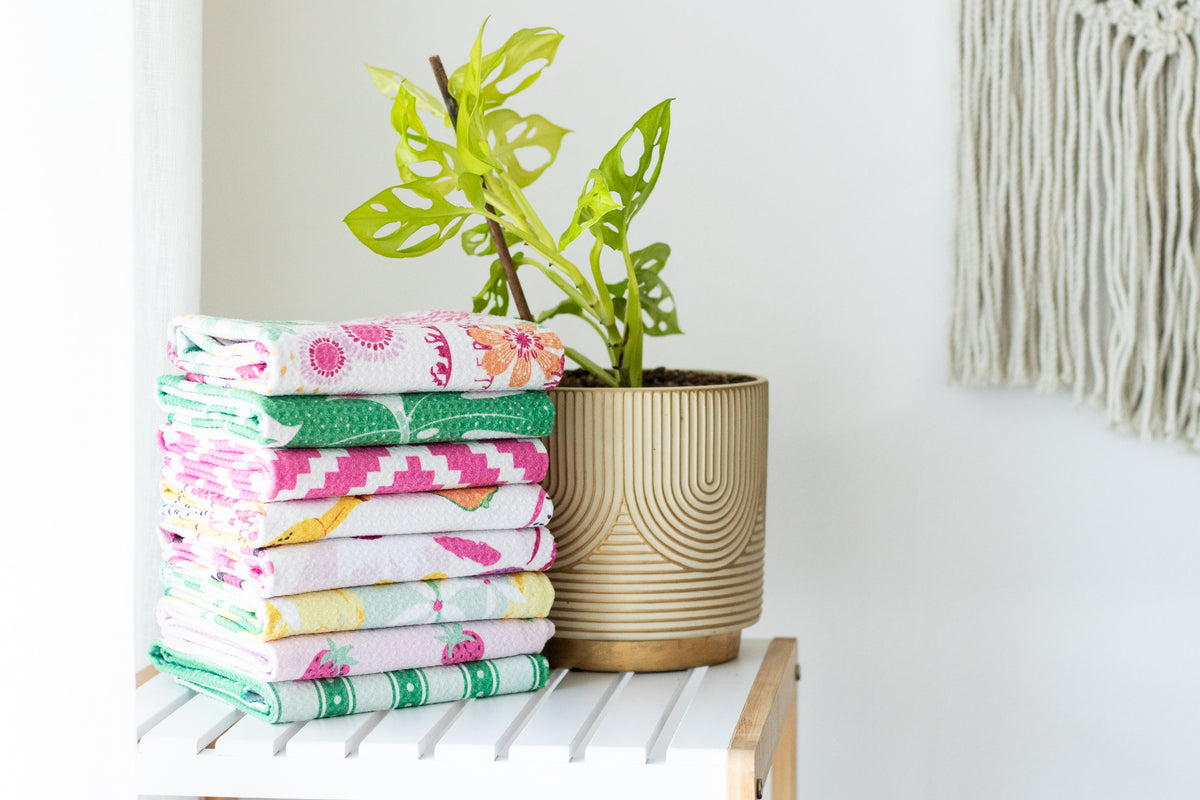 A stack of folded waffle-weave kitchen tea towels sits on a white slatted side table next to a potted green plant in a ribbed beige pot, against a white wall.