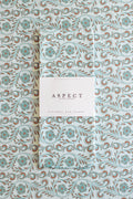A folded waffle-weave kitchen tea towel with a repeating floral pattern in light blue, turquoise, and brown, wrapped with a white paper belly band label that reads 'ASPECT HOME GOODS' and "KITCHEN TEA TOWEL', laid flat on a towel with the same pattern.
