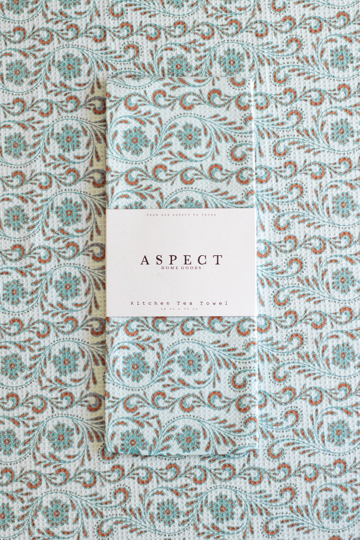 A folded waffle-weave kitchen tea towel with a repeating floral pattern in light blue, turquoise, and brown, wrapped with a white paper belly band label that reads 'ASPECT HOME GOODS' and "KITCHEN TEA TOWEL', laid flat on a towel with the same pattern.