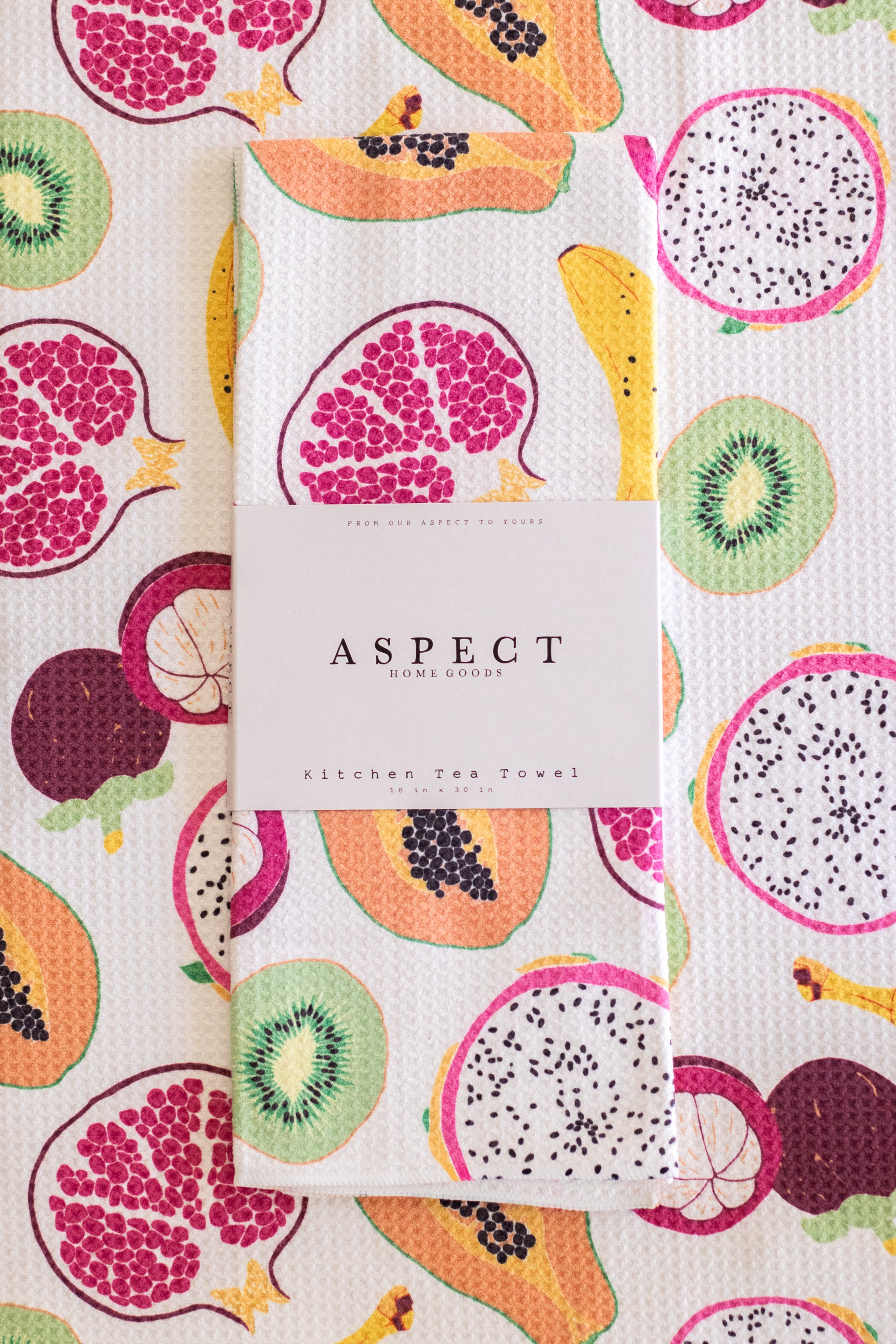 A folded kitchen tea towel with a colorful tropical fruit print is displayed on top of a matching flat tea towel. A white paper belly band around the folded towel reads 'ASPECT HOME GOODS' and 'KITCHEN TEA TOWEL'.