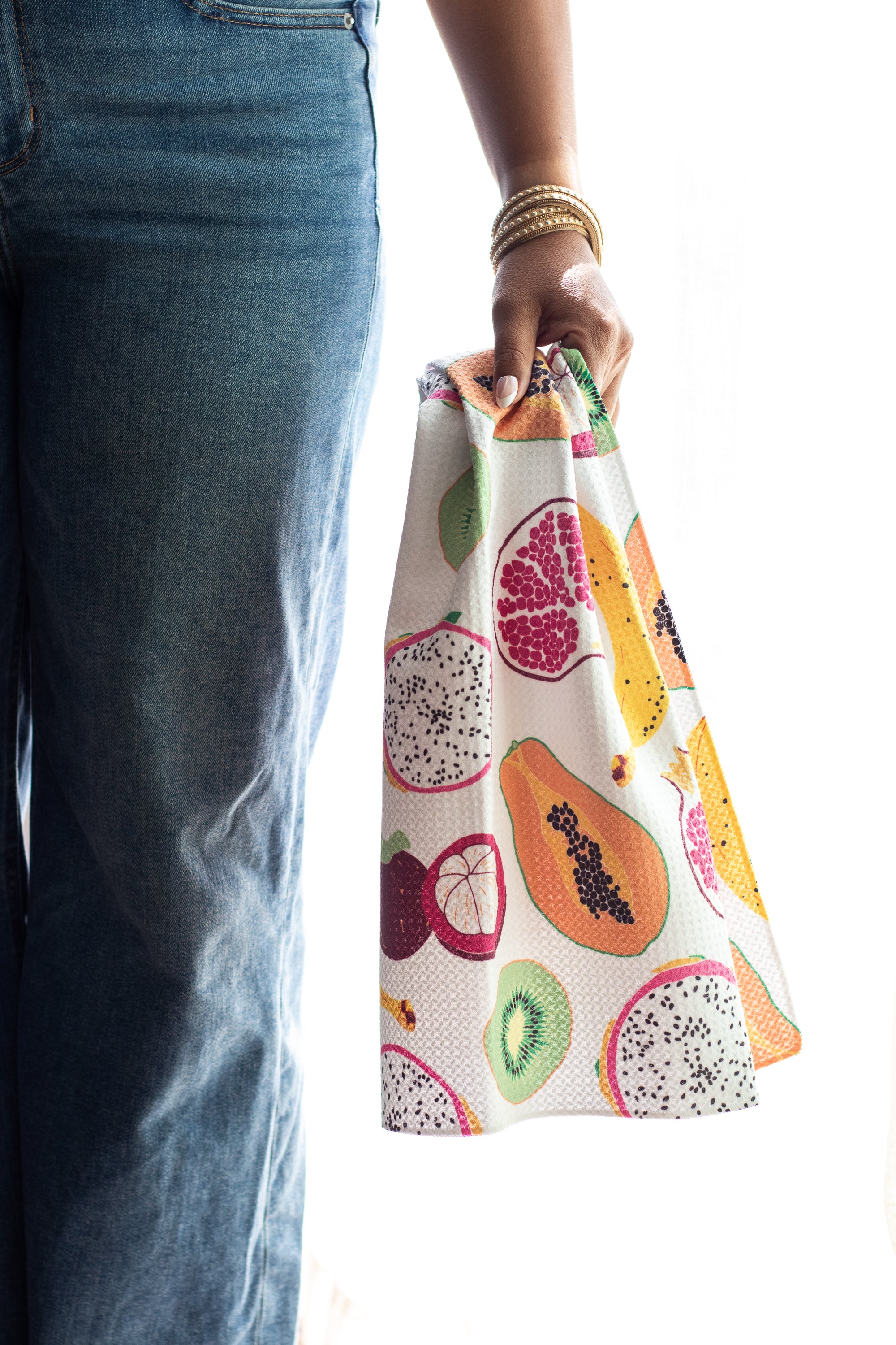 A person wearing blue jeans holds a waffle-weave kitchen tea towel printed with colorful tropical fruit illustrations, including papayas, dragon fruit, kiwi, bananas, pomegranate, and mangosteen, against a bright white background.