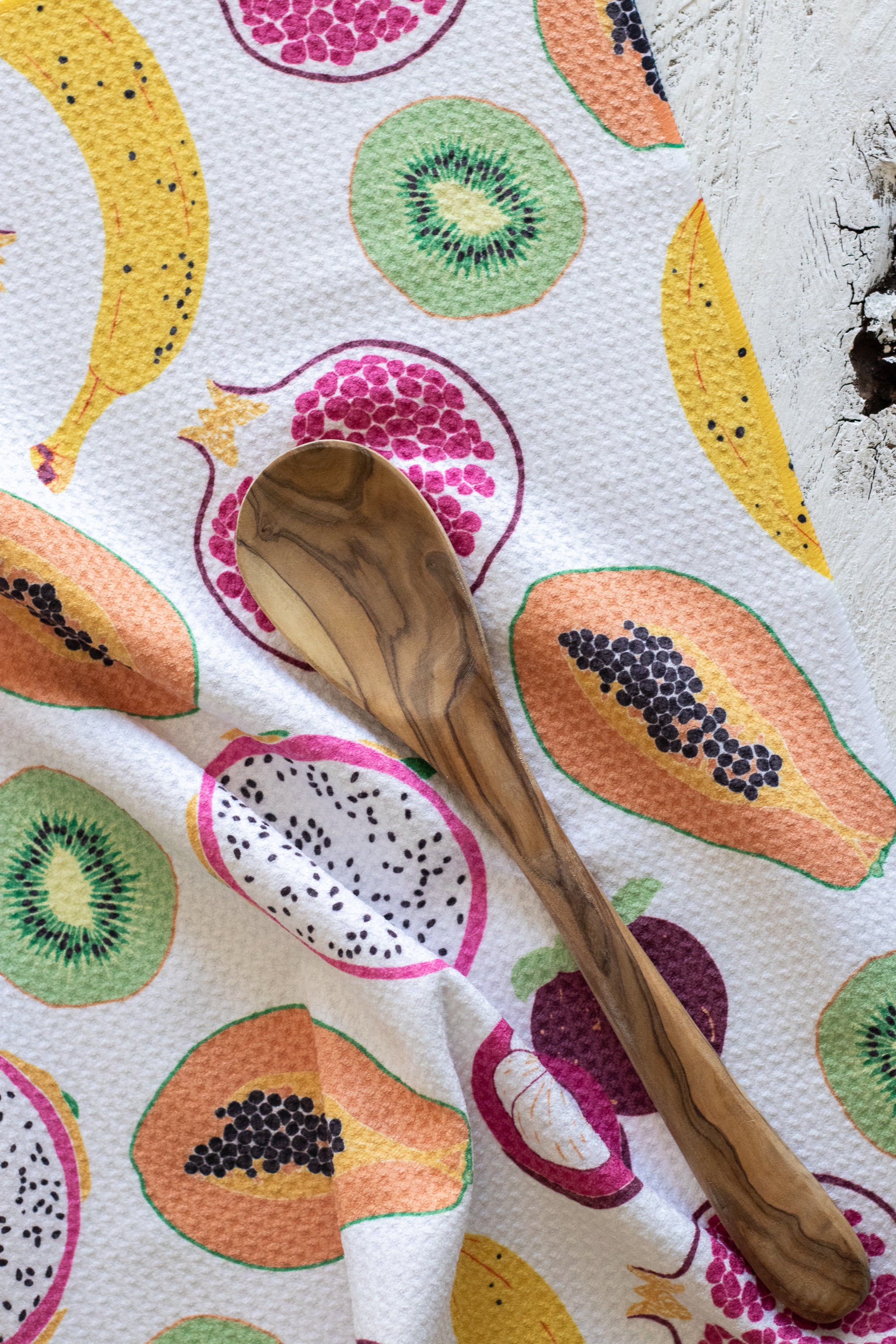 A wooden spoon with a dark olive-wood grain pattern rests on a white waffle-weave kitchen towel printed with colorful illustrations of tropical fruits including papayas, mangosteen, pomegranate, kiwi, dragon fruit, and bananas.