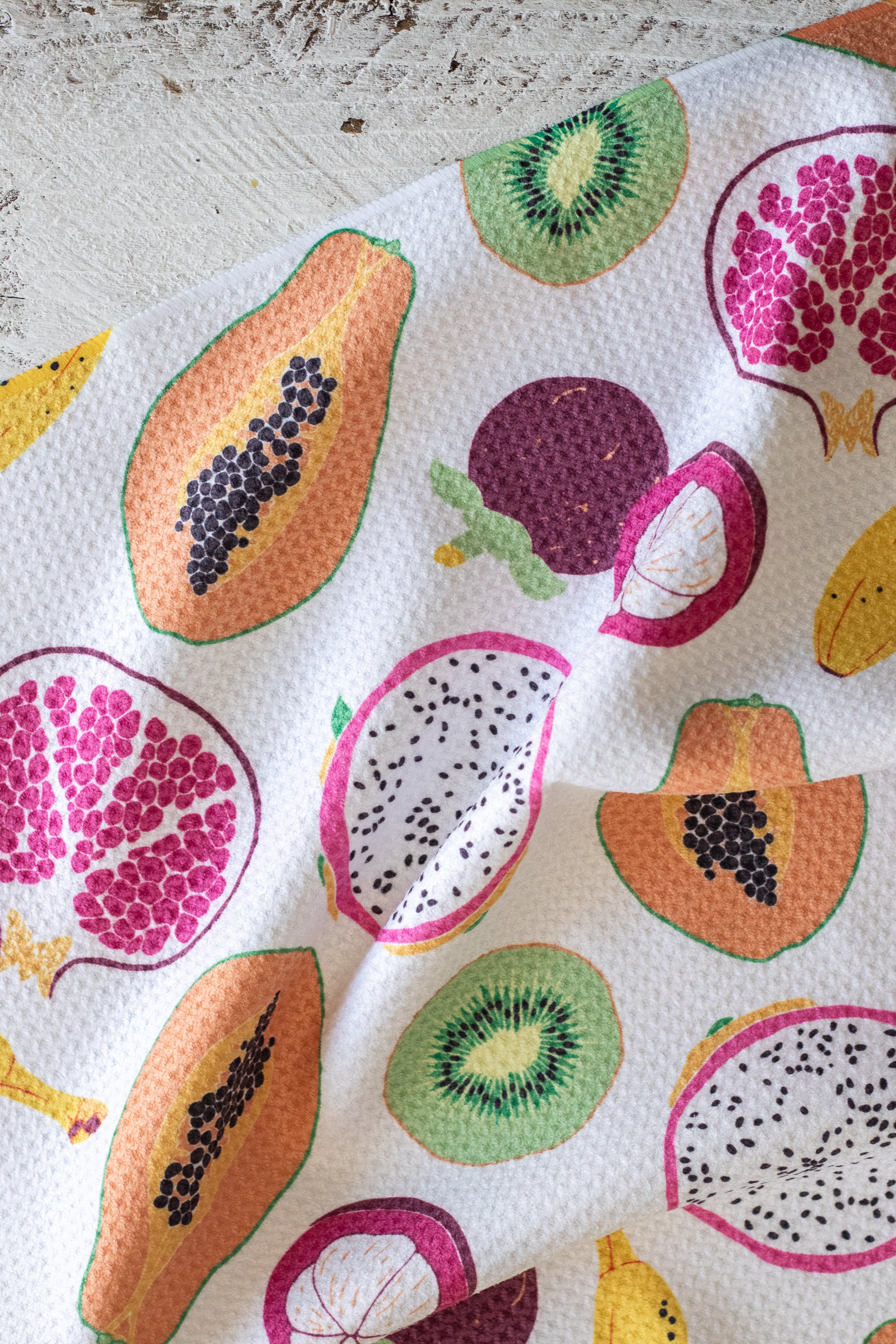 A close-up of a waffle-weave kitchen tea towel or napkin, printed with a colorful pattern of sliced tropical fruits including papayas, mangosteen, bananas, dragon fruit, kiwi, and pomegranate.