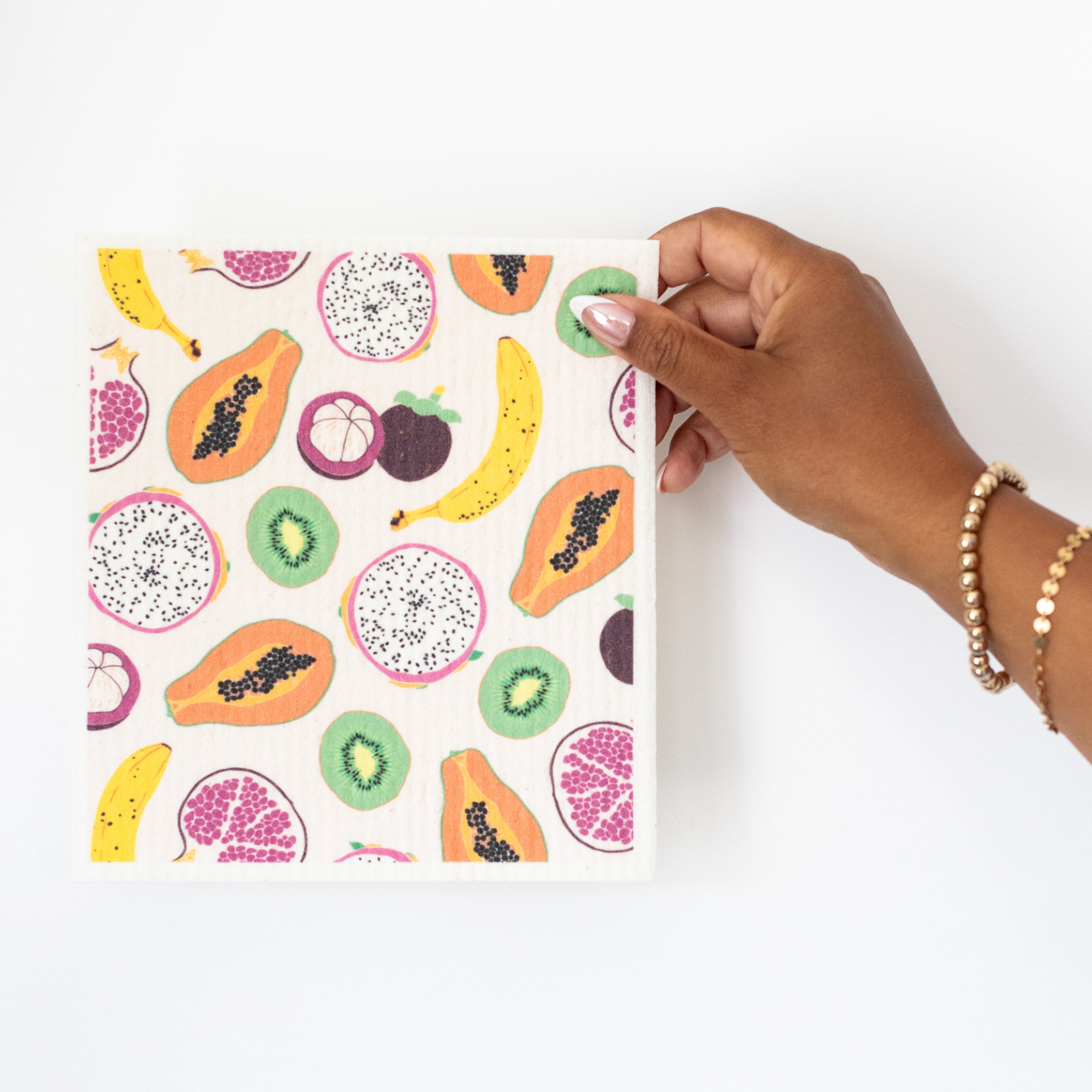 A hand holding a Swedish dishcloth with a colorful tropical fruit pattern that includes bananas, kiwi, dragonfruit, papayas, and mangosteen, on a white background.