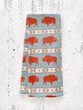 A waffle-weave kitchen tea towel with western-style terracotta-colored bison design on a light blue background with beige southwestern geometric pattern stripes, folded lengthwise in half, on a textured white surface.