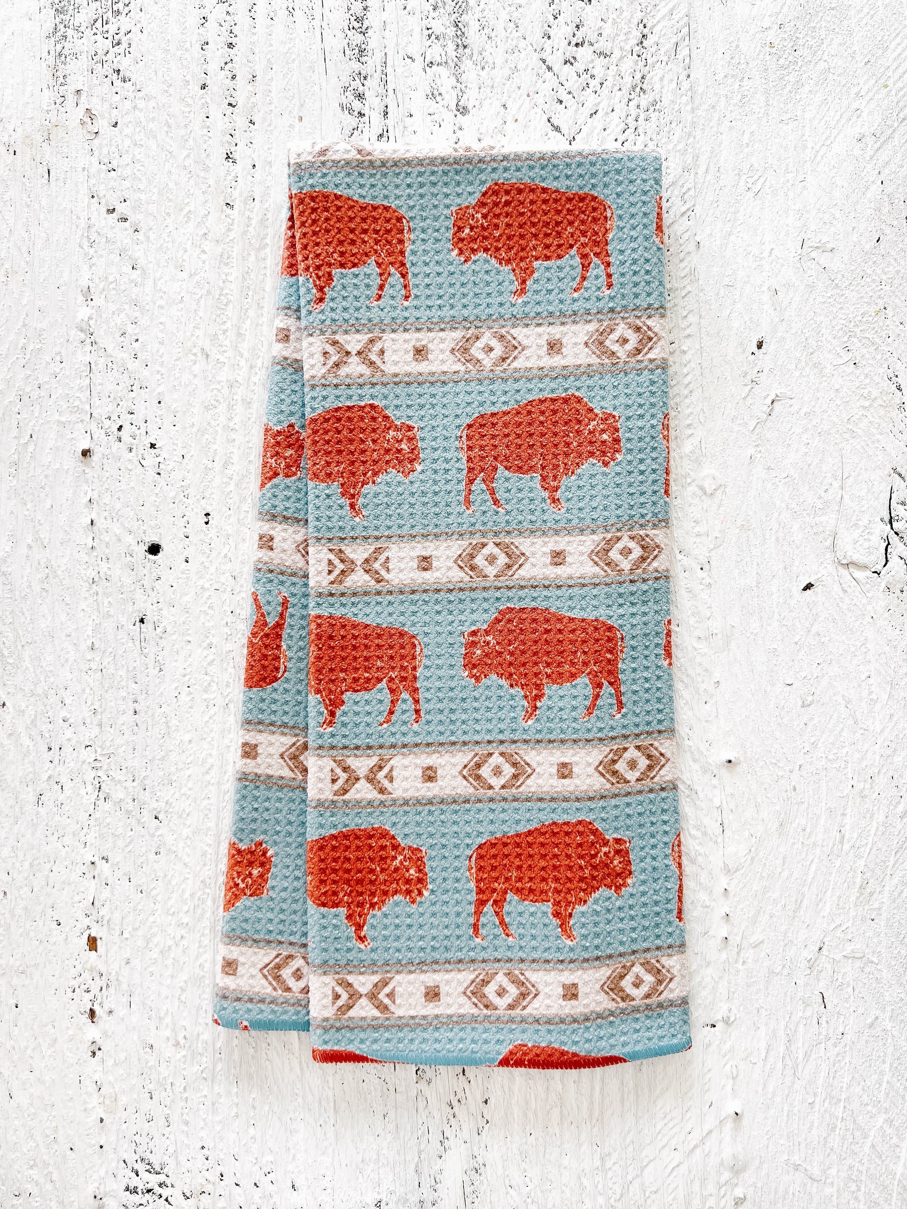 A waffle-weave kitchen tea towel with western-style terracotta-colored bison design on a light blue background with beige southwestern geometric pattern stripes, folded lengthwise in half, on a textured white surface.