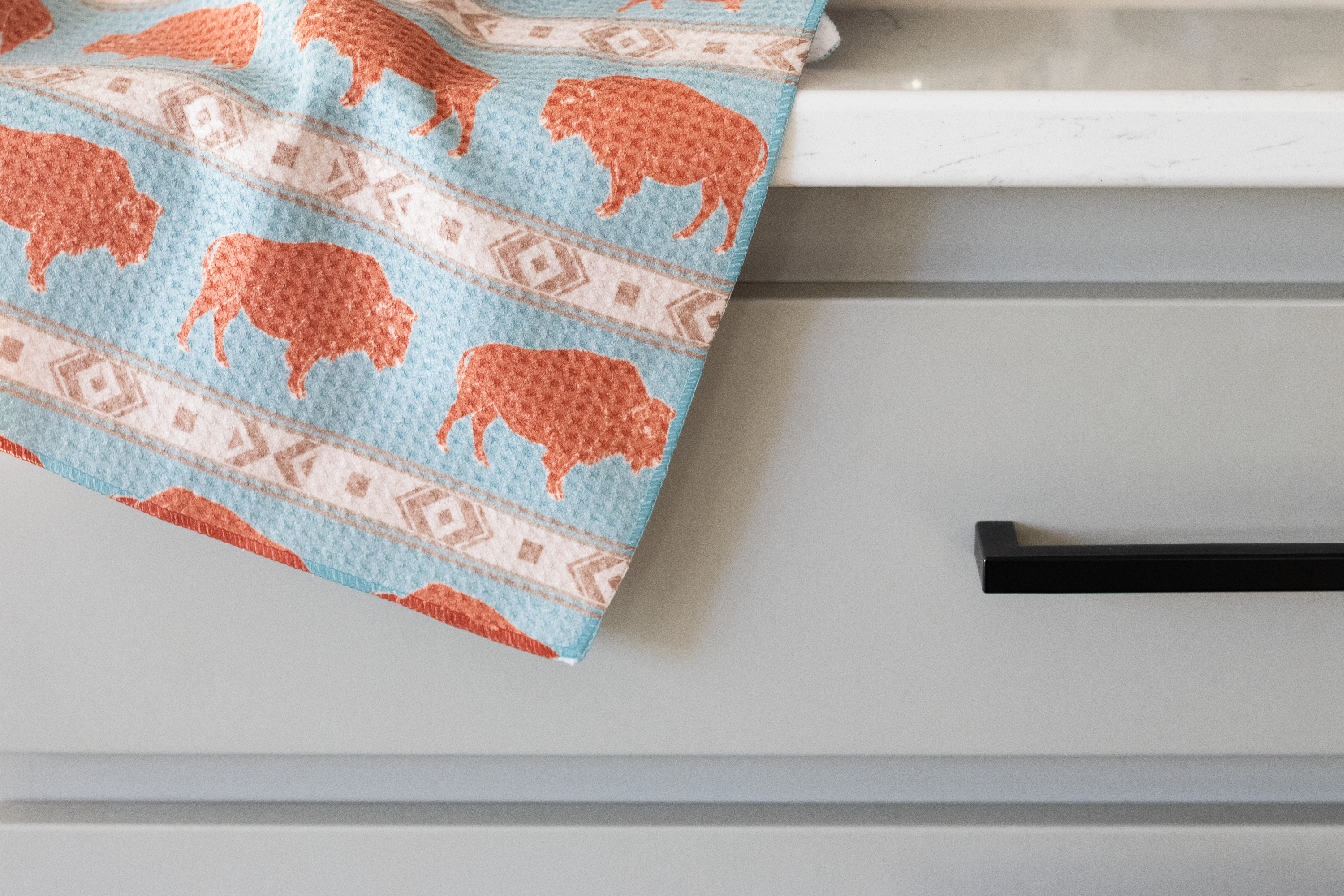 Patterned waffle-weave kitchen tea towel with a terracotta-colored bison and beige southwestern geometric stripe designs on a light blue background, draped over the edge of a white counter in front of a gray drawer with a black handle.