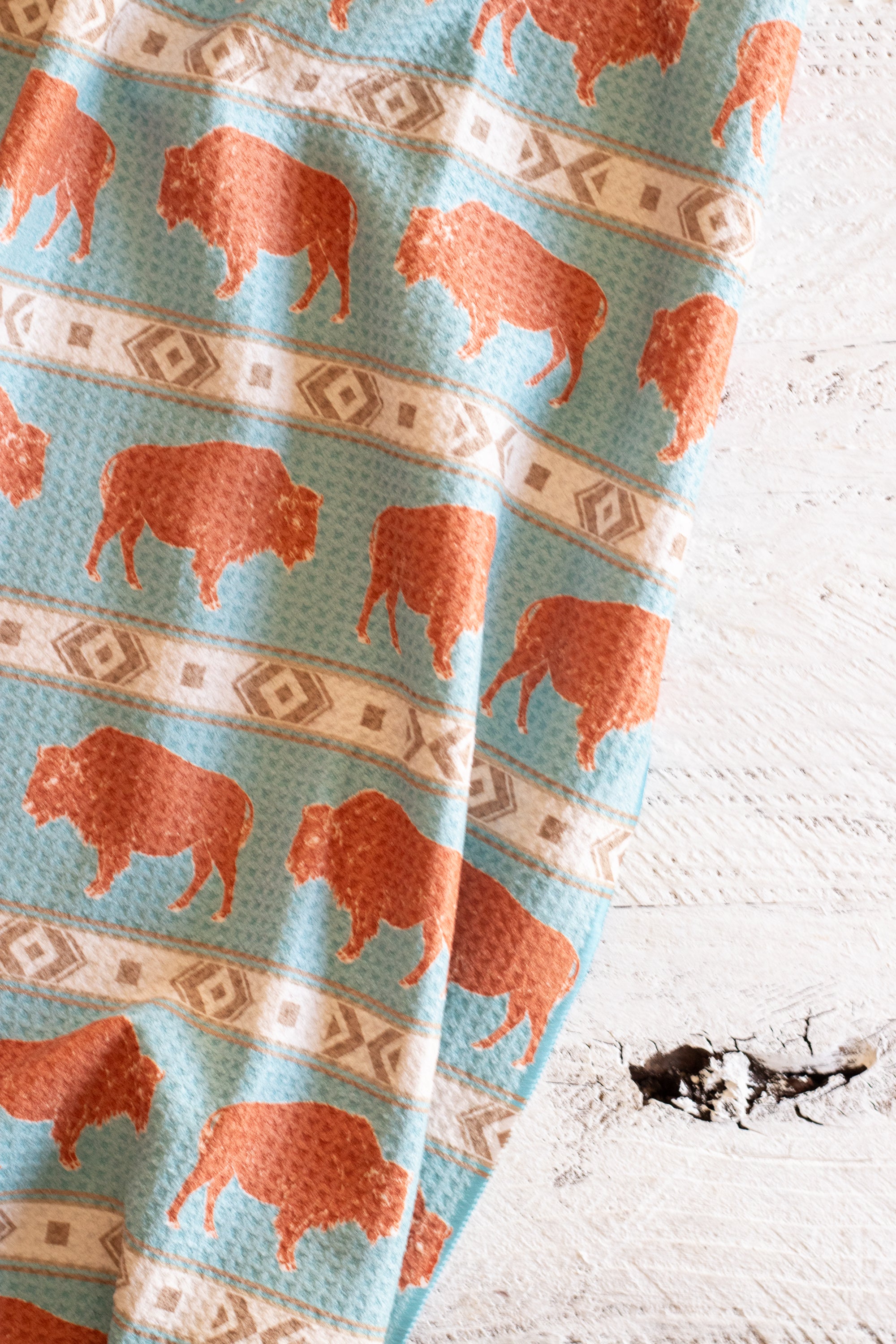 A patterned waffle-weave kitchen tea towel with a terracotta-colored bison and beige southwestern geometric stripe design on a light blue background, on a white rustic wooden surface.