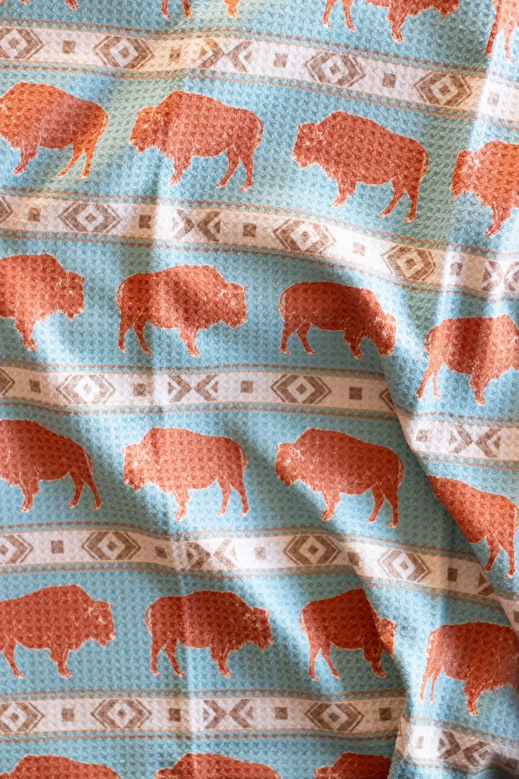 A blue waffle-weave kitchen tea towel with a pattern of terracotta-colored bison and horizontal beige geometric stripes.