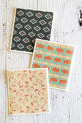 Three Swedish dishcloths with Western-style designs are arranged on a distressed white wooden surface. The dishcloth at the top has a black background with a repeating turquoise, red, and white geometric diamond pattern, the middle one has a horizontal geometric stripe pattern with bison in turquoise and terracotta, and the bottom dishcloth has a horse pattern in red and beige.