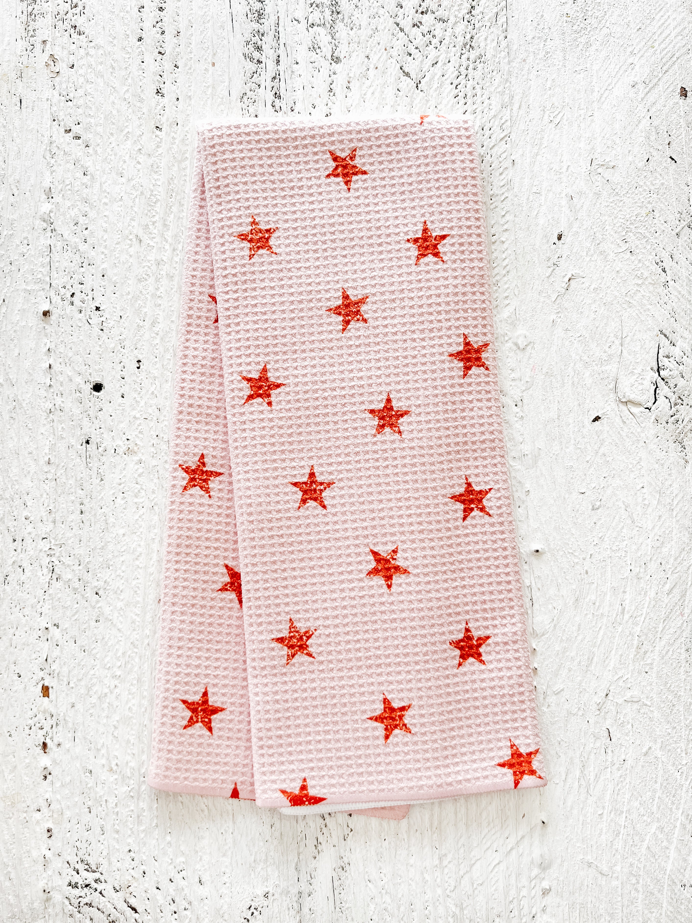 A waffle-weave kitchen tea towel with a pattern of terracotta-colored, distressed stars on a light peachy-pink background, folded lengthwise in half, on a textured white surface.