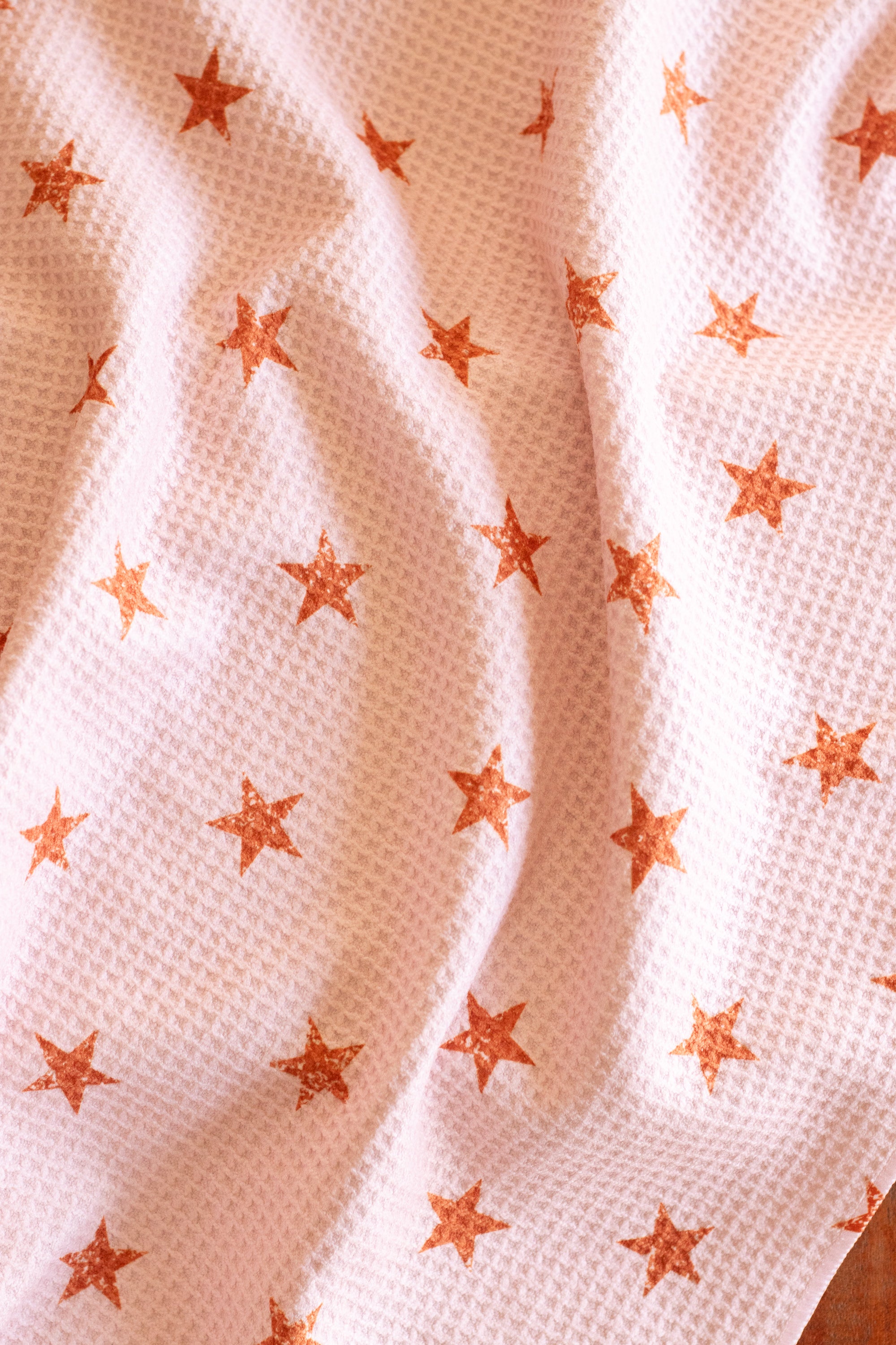 Close-up of a waffle-weave kitchen tea towel with a distressed terracotta-colored star pattern on a light peachy-pink background.