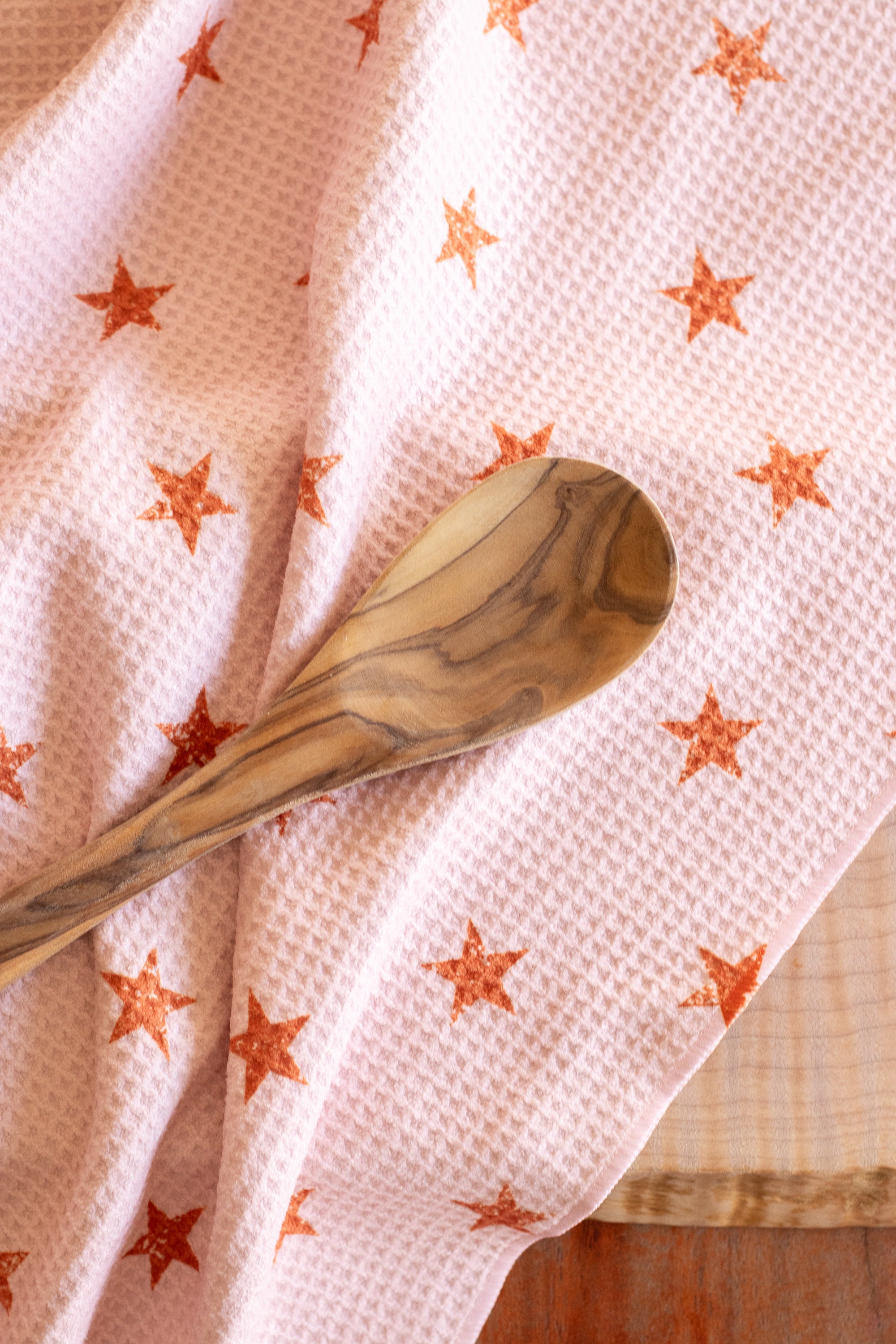 Close-up of a wooden spoon on a waffle-weave kitchen tea towel with a distressed terracotta-colored star pattern on a light peachy-pink background, on a wooden surface.