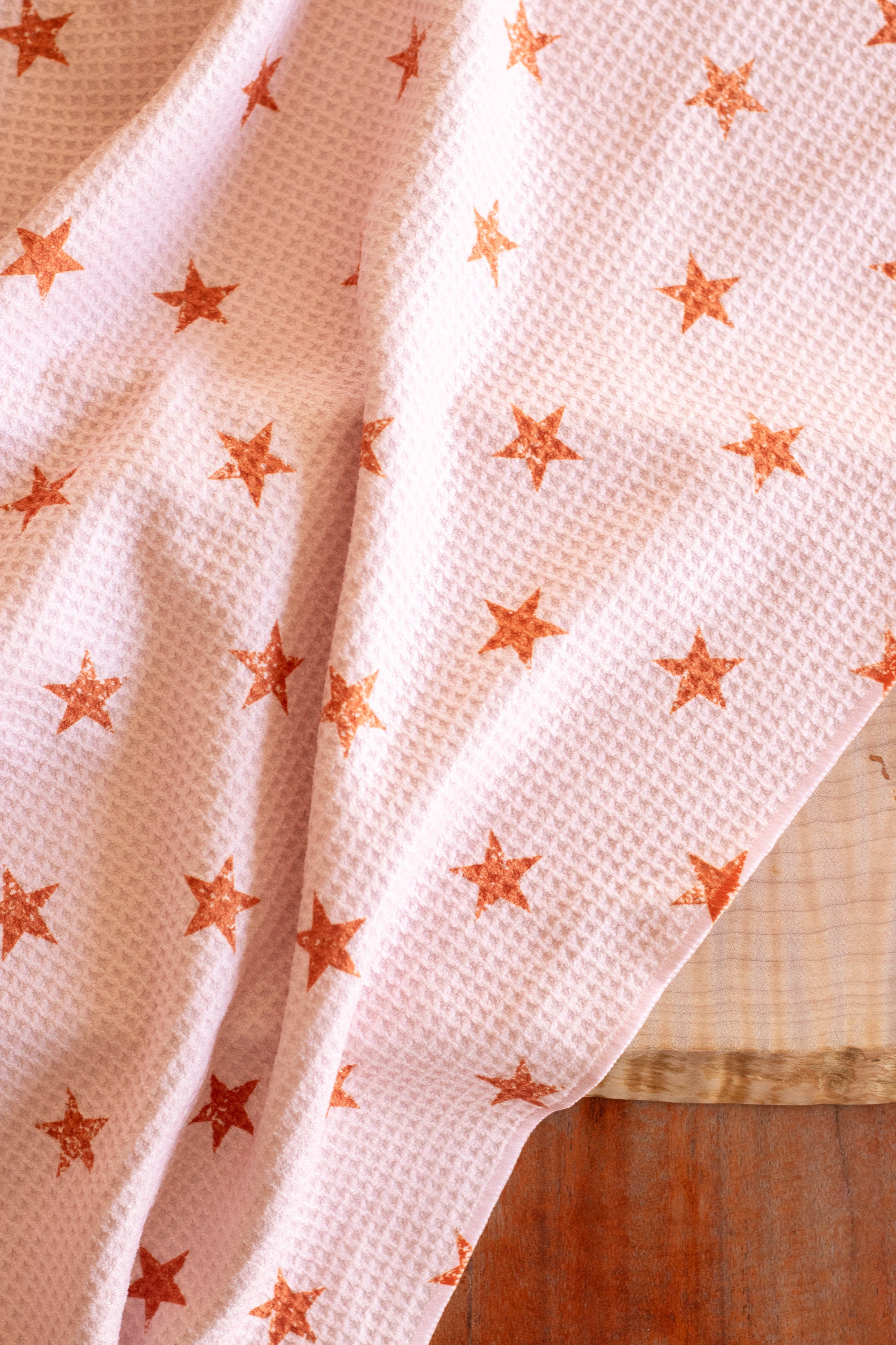 Close-up of a waffle-weave kitchen tea towel with a distressed terracotta-colored star pattern on a light peachy-pink background, on a wood surface.