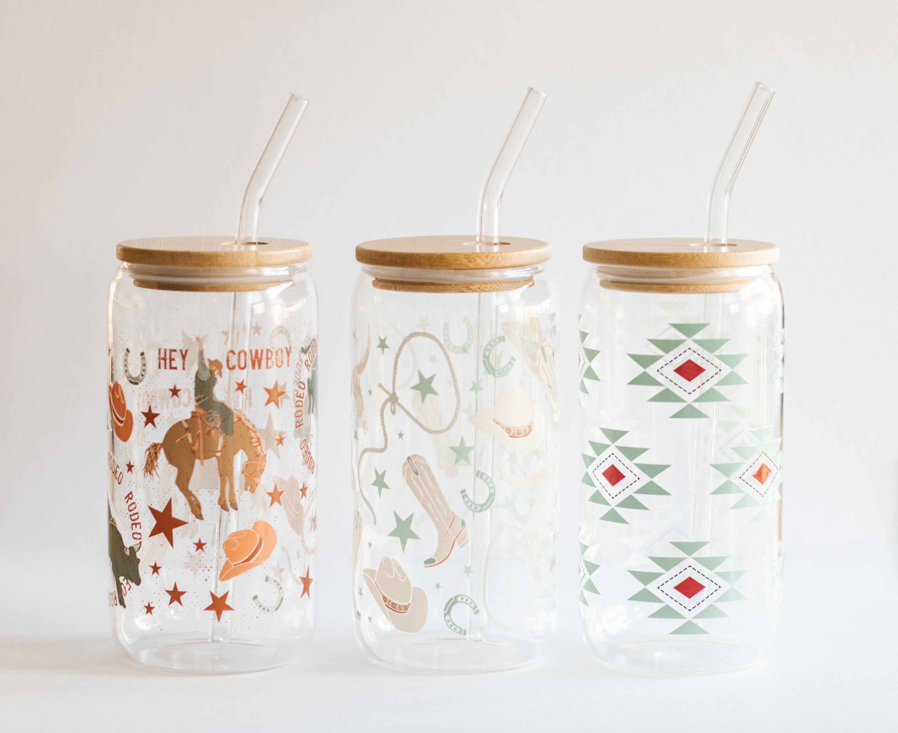 Three can glass tumblers with bamboo wooden lids and glass straws, featuring cowboy-themed designs on a white background.