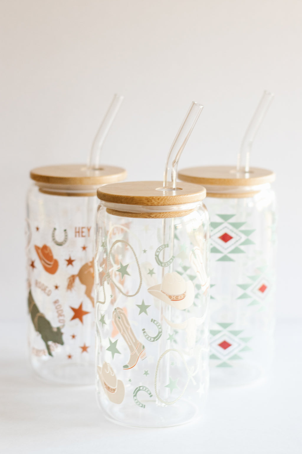 Three can glass tumblers with bamboo lids and glass straws featuring different western-style designs on a white background