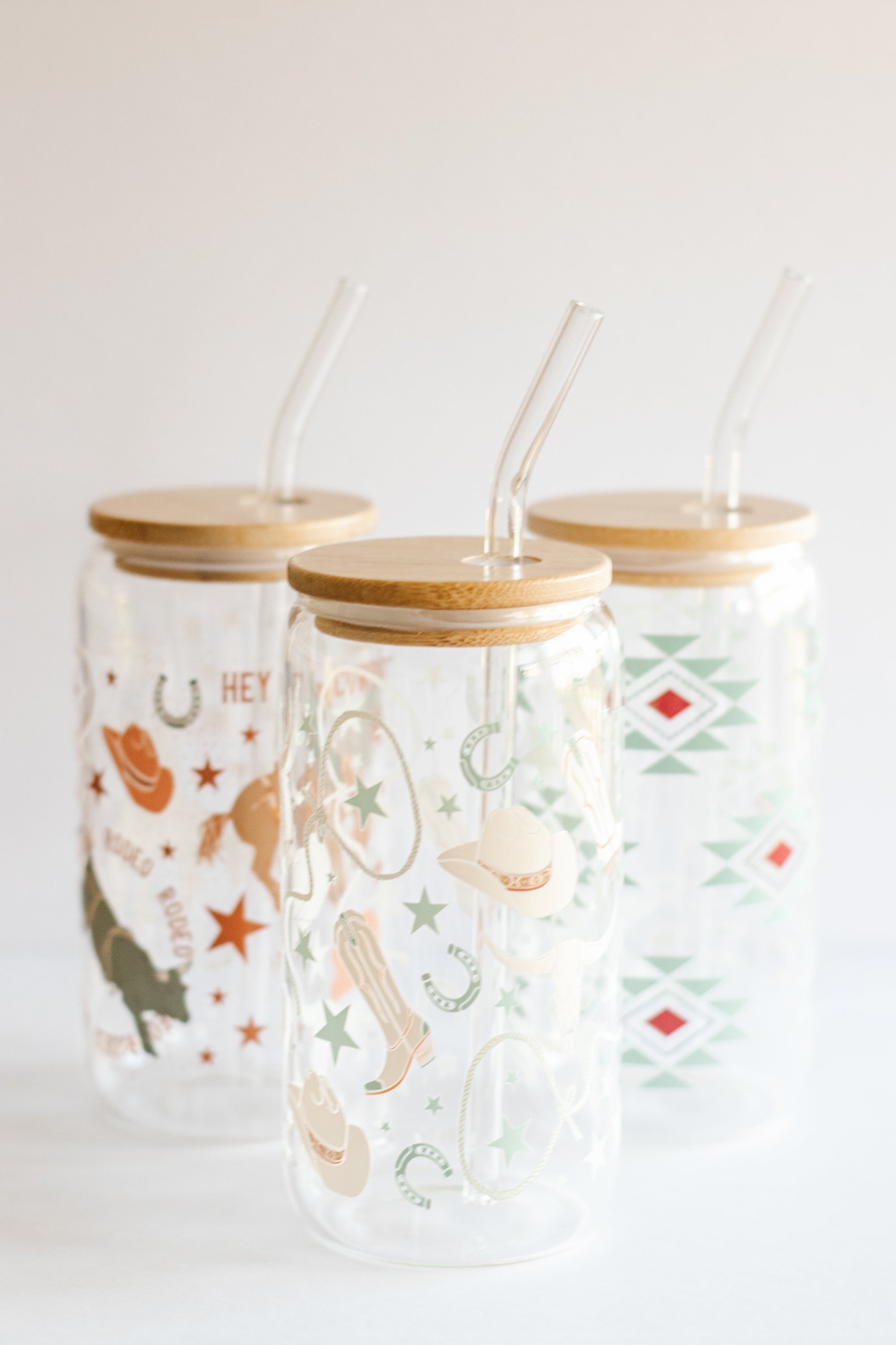 Three can glass tumblers with bamboo lids and glass straws featuring different western-style designs on a white background