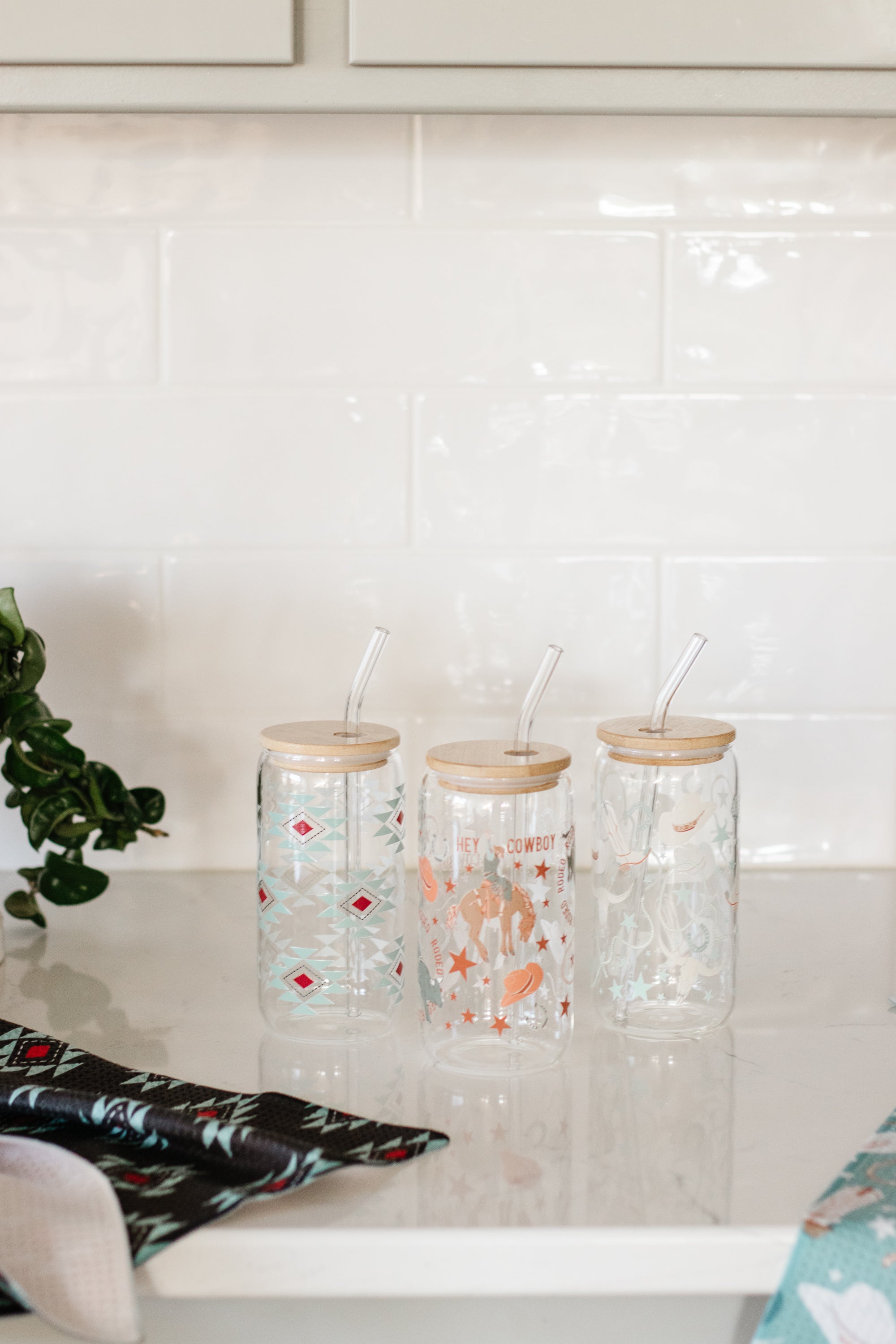 Three can glasses featuring western-style designs with bamboo wooden lids and glass straws on a kitchen counter.
