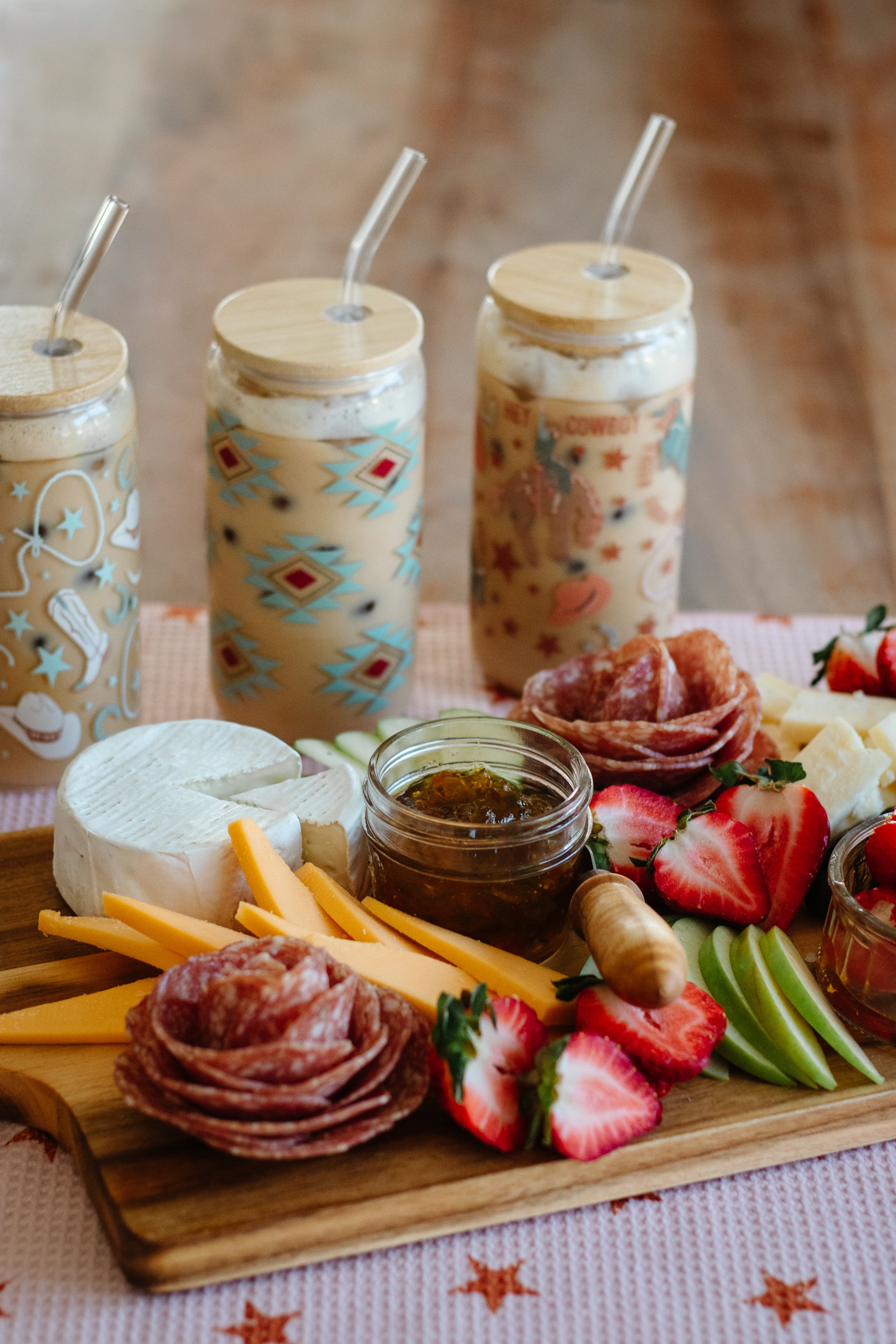 Platter of assorted meats, cheeses, fruits, and a jar of jam on a star-patterned pink towel with western-style can glasses in the background.