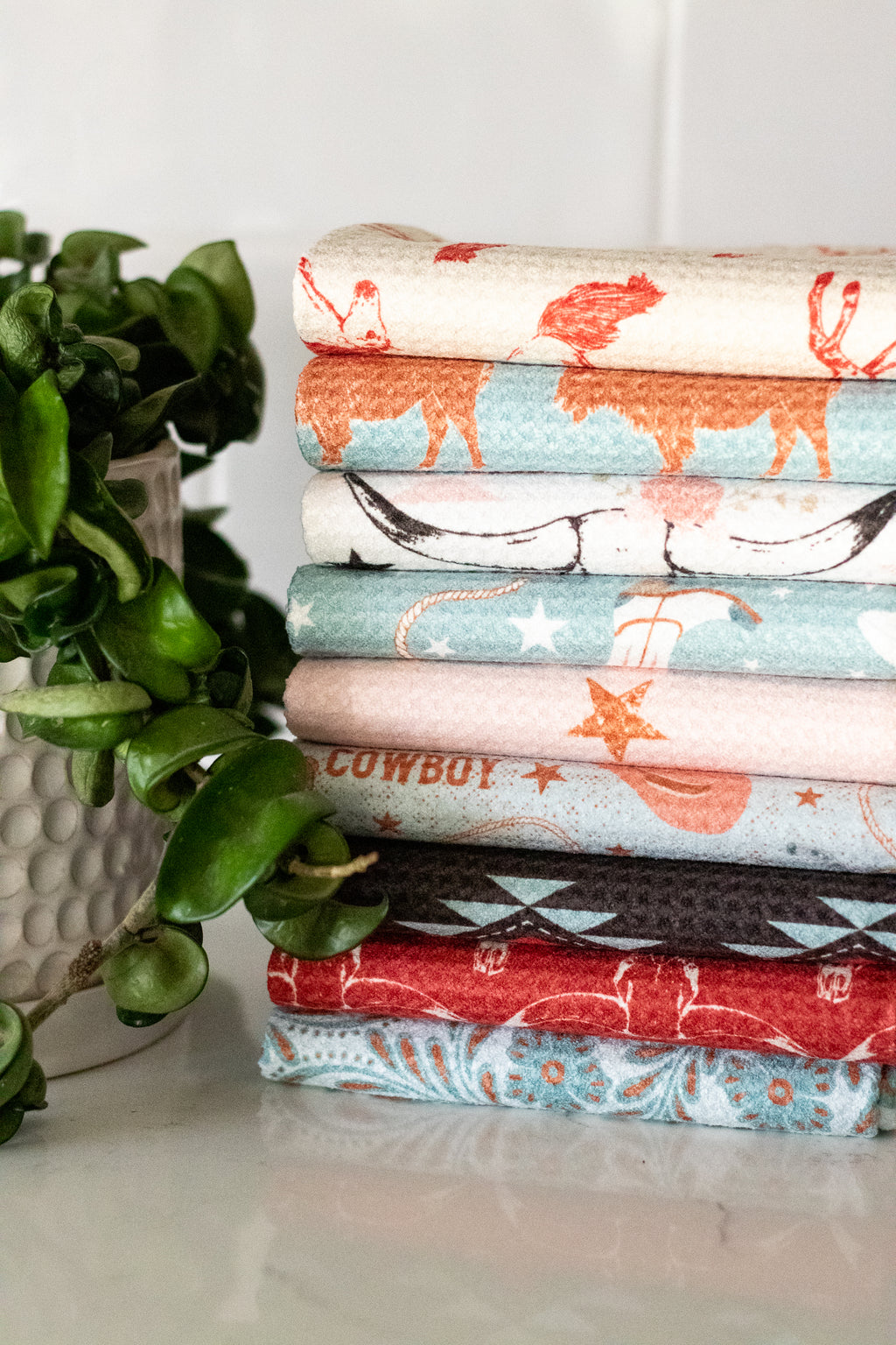 Stack of folded waffle-weave kitchen tea towels with various western patterns and designs on a white surface, next to a plant.
