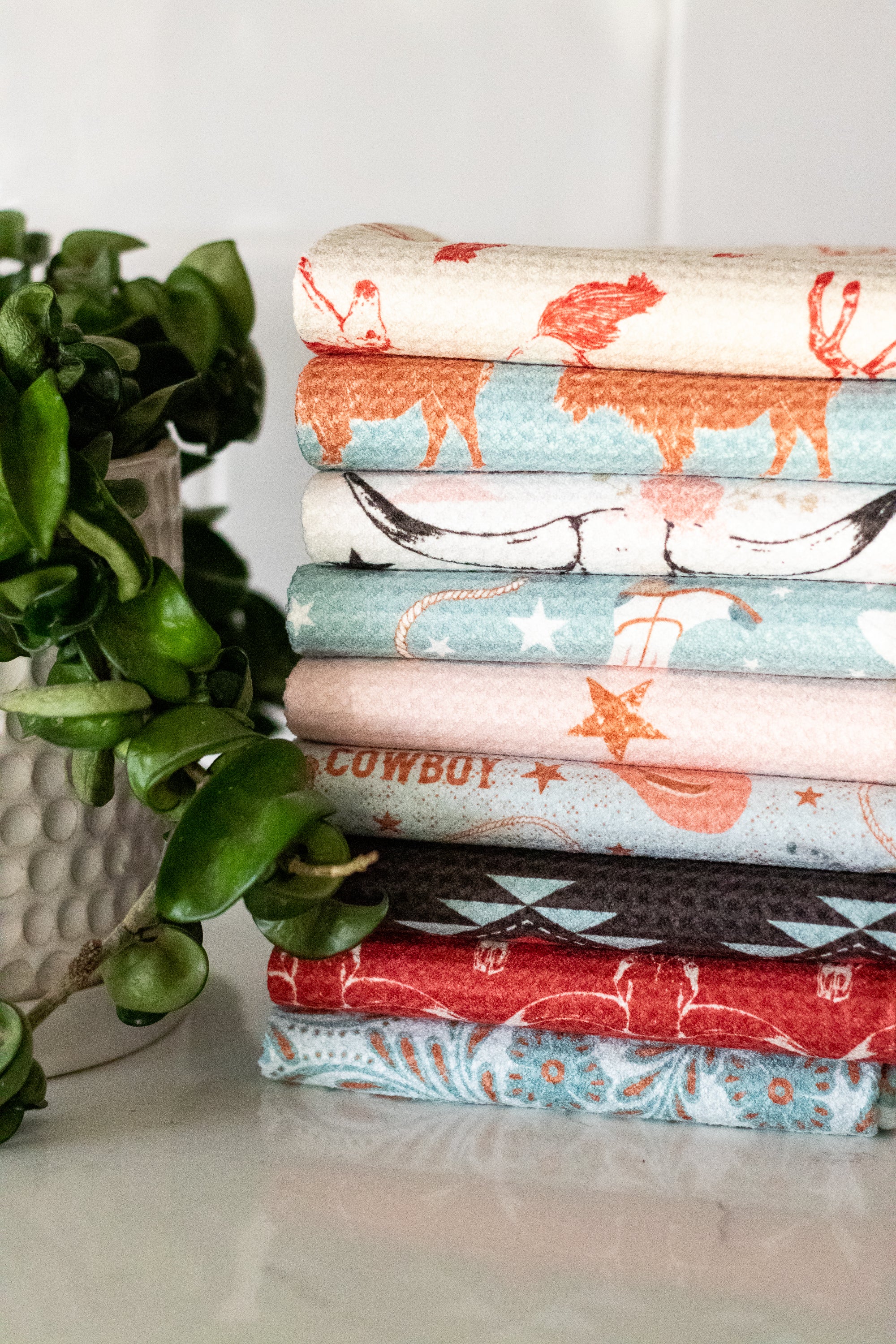 Stack of folded waffle-weave kitchen tea towels with various western patterns and designs on a white surface, next to a plant.