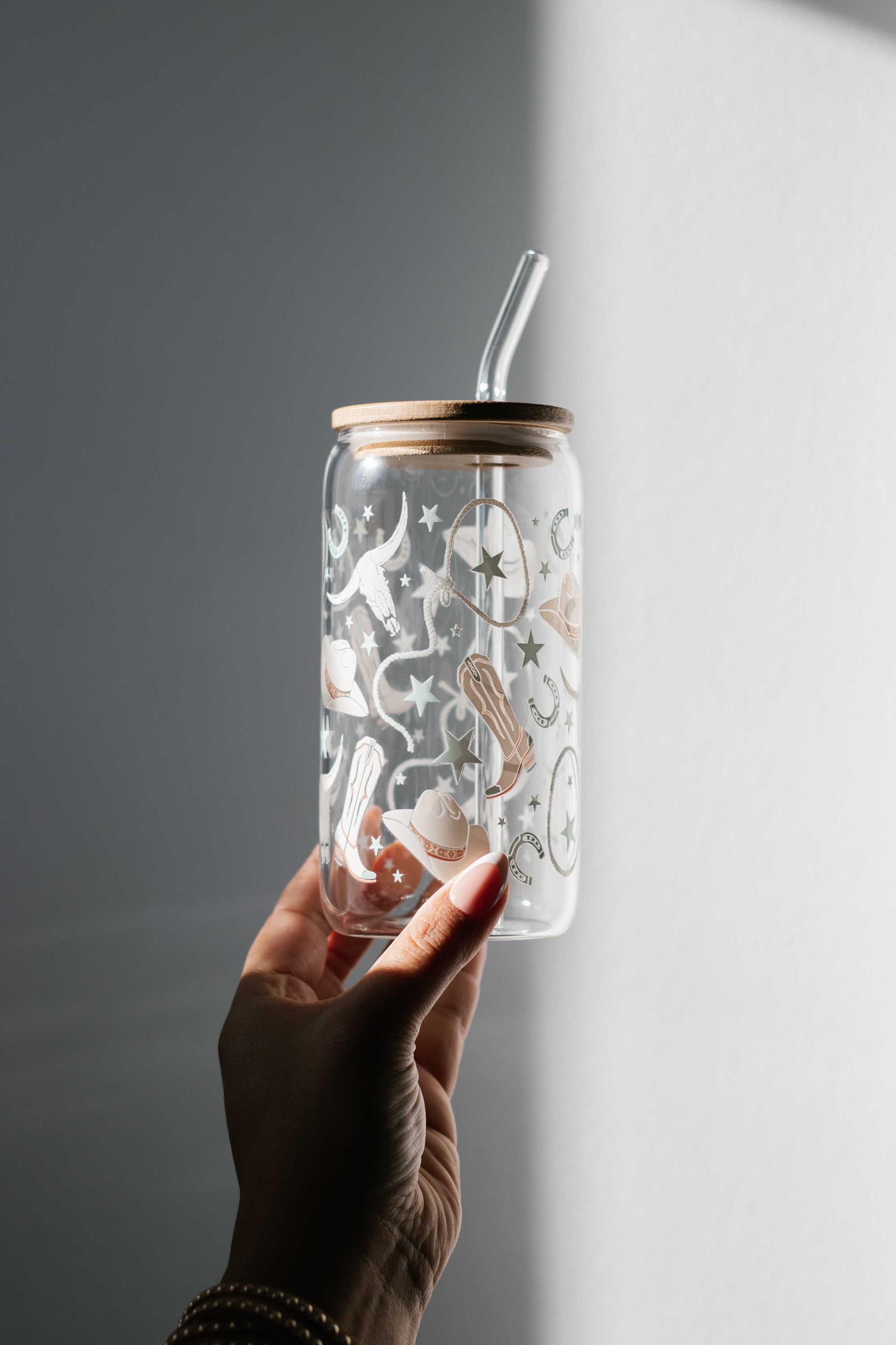 Hand holding a can glass tumbler with a bamboo lid and a glass straw against a gray background