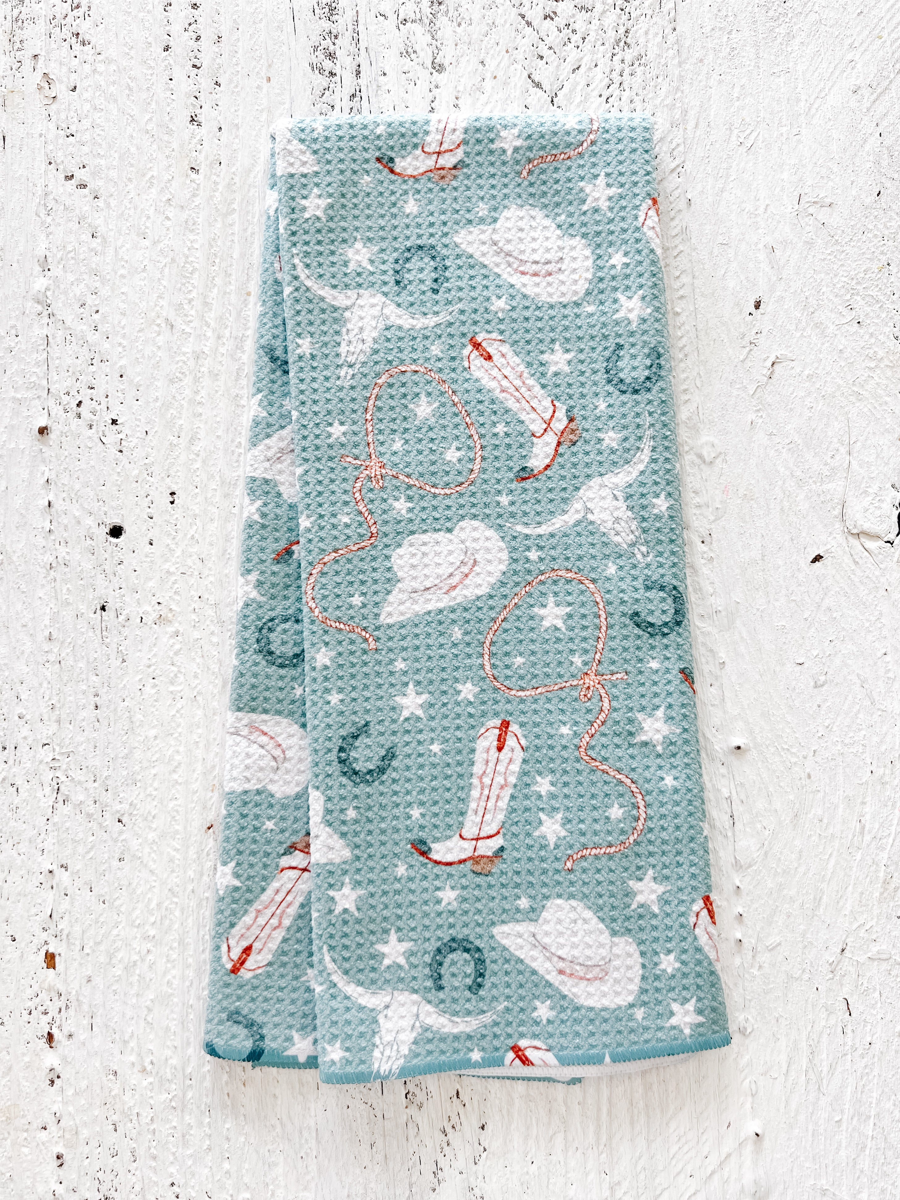 A blue waffle-Weave kitchen tea towel with a cowgirl pattern, including lassos, hats, horseshoes, cow skulls, and stars, on a textured white background.