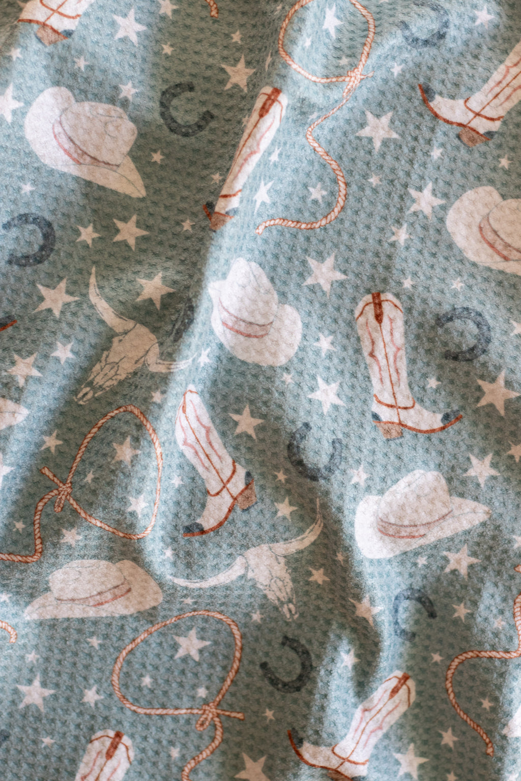Close-up of a waffle-weave kitchen tea towel featuring a pattern of cowgirl hats, boots, stars, lassos, and horseshoes.