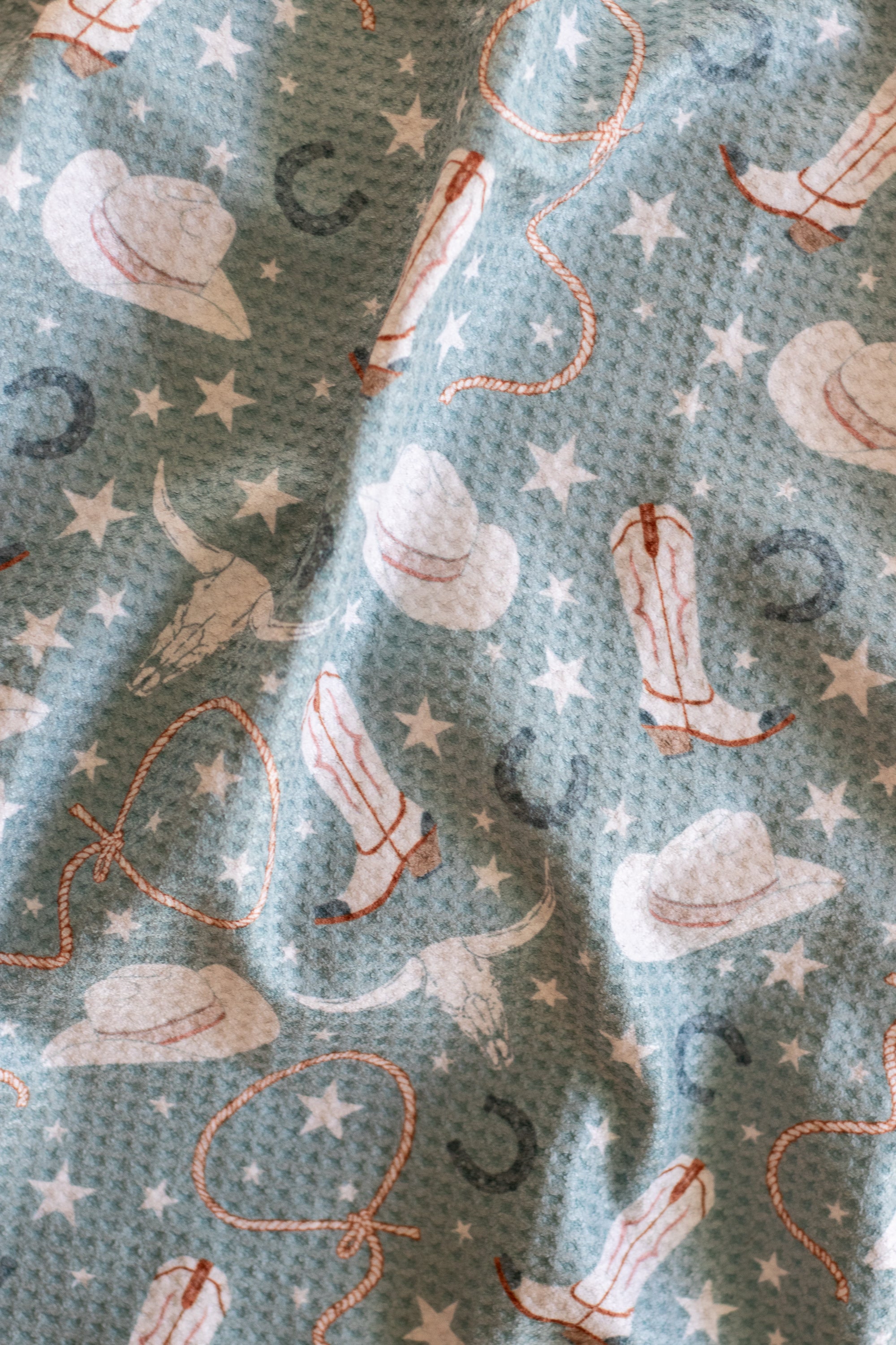 Close-up of a waffle-weave kitchen tea towel featuring a pattern of cowgirl hats, boots, stars, lassos, and horseshoes.