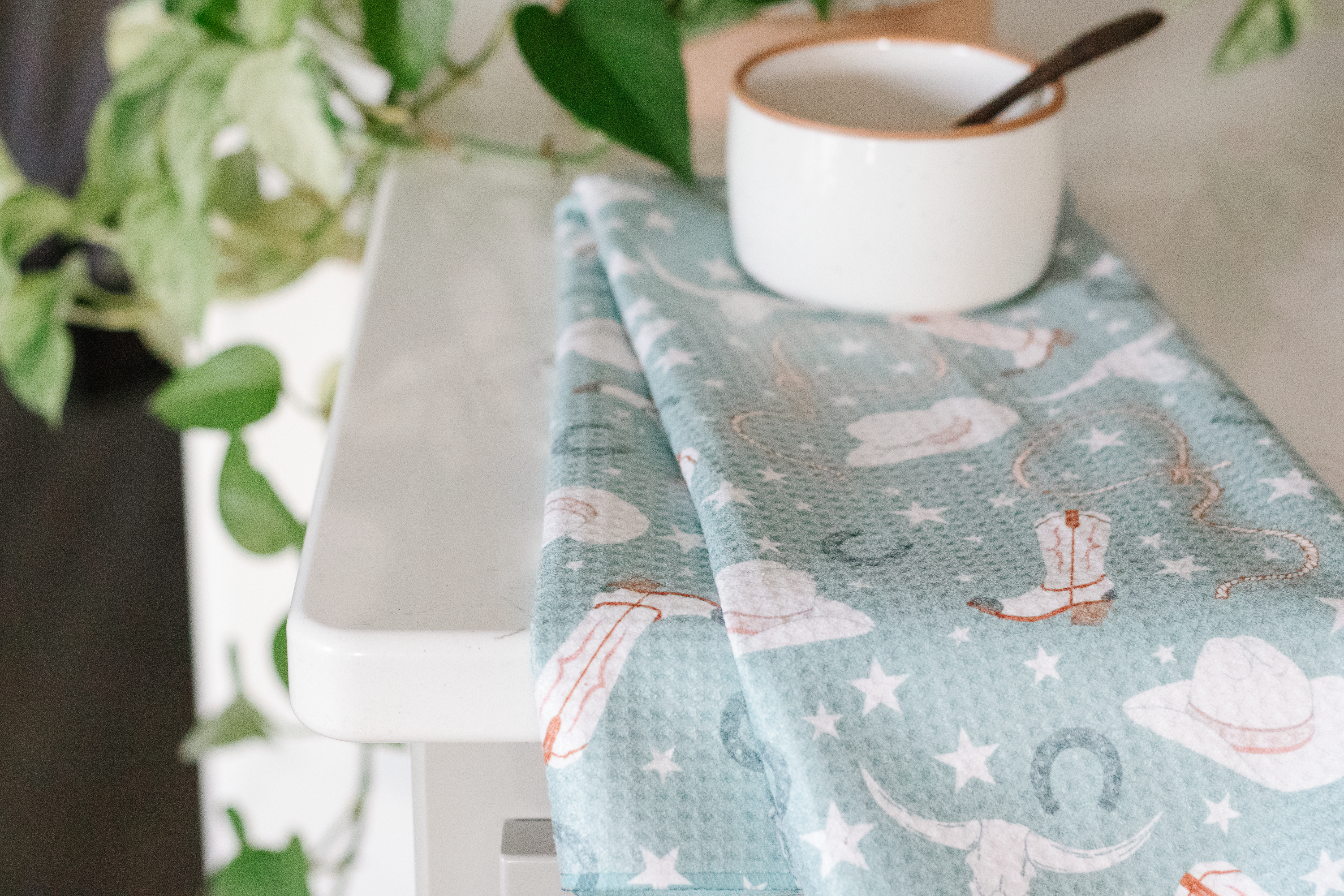 Waffle-weave tea towel with a cowgirl pattern on a kitchen counter with a ramekin and spoon.