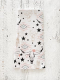 A waffle-weave kitchen tea towel with a western-style pattern of hand-drawn longhorn cow skulls, cacti, stars, and southwestern geometric shapes in muted colors of pinks, beiges, and black, with a white background, folded lengthwise in half, on a textured white surface.