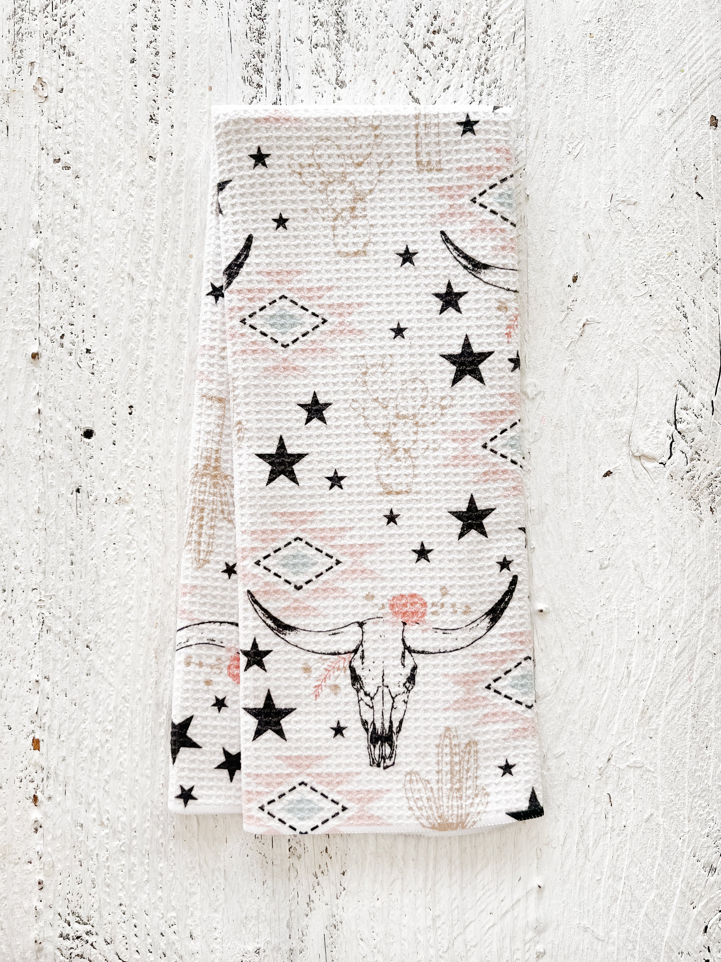 A waffle-weave kitchen tea towel with a western-style pattern of hand-drawn longhorn cow skulls, cacti, stars, and southwestern geometric shapes in muted colors of pinks, beiges, and black, with a white background, folded lengthwise in half, on a textured white surface.