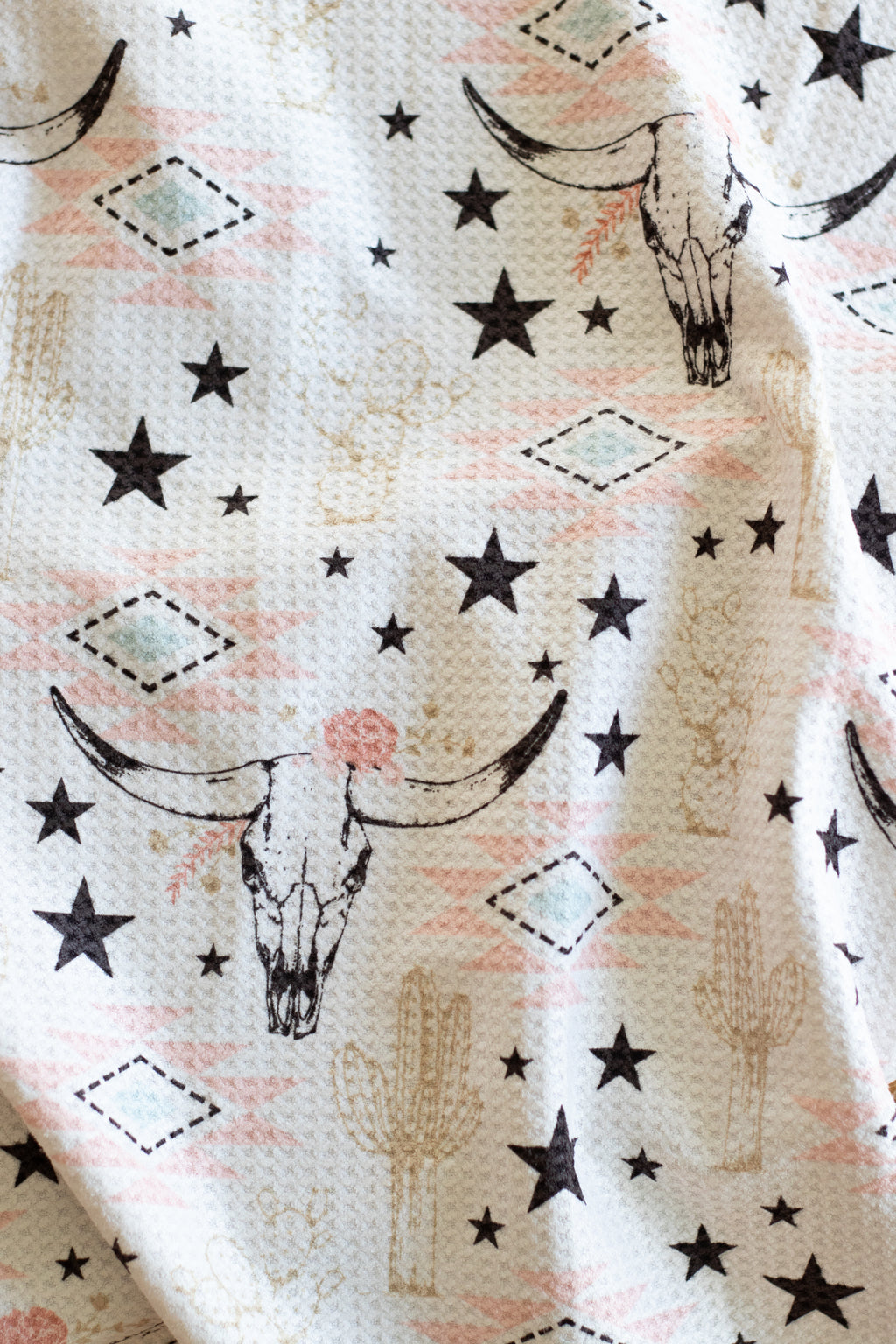 A waffle-weave kitchen tea towel with a longhorn cow skull, stars, and cactus pattern in muted pinks, beige, blue, and black, on a white background.