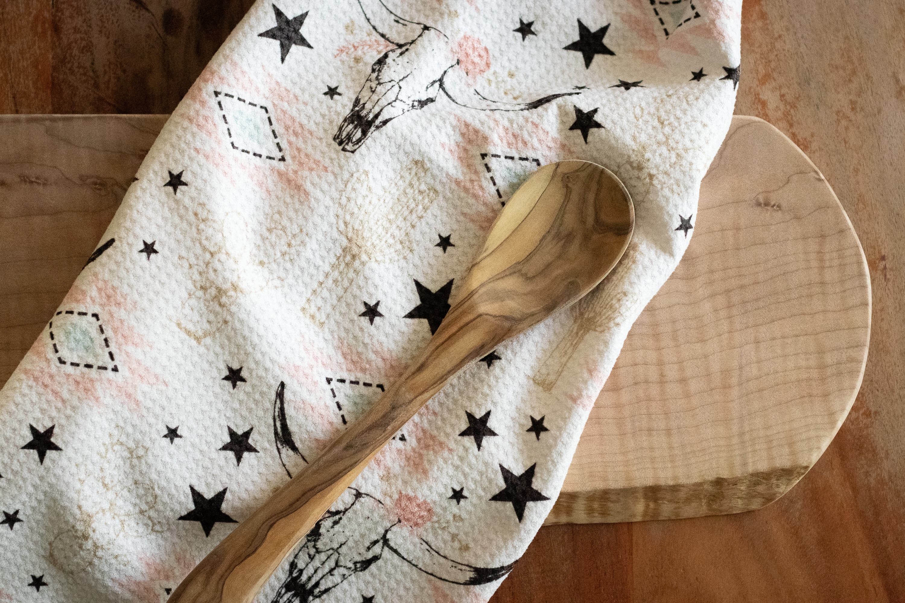 A wooden spoon on a waffle-weave tea towel with a longhorn cow skull, stars, and cactus pattern in muted pinks, beige, blue, and black, on a white background, lying on a wooden surface.