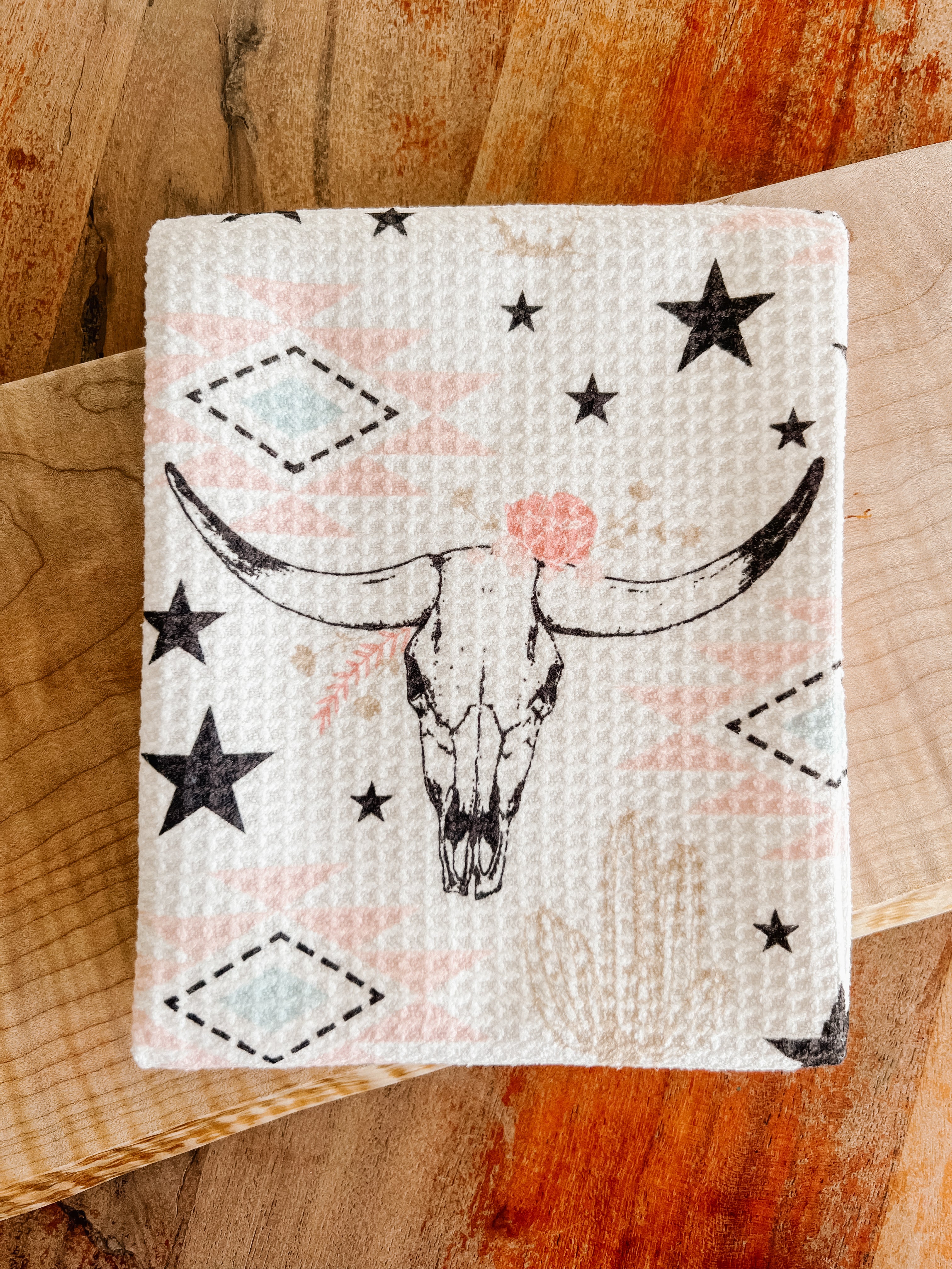 A folded waffle-weave tea towel with a pattern of longhorn cow skulls, stars, cacti, and southwestern geometric shapes, in muted pinks, beige, blue, and black, on a white background, on a wood surface.
