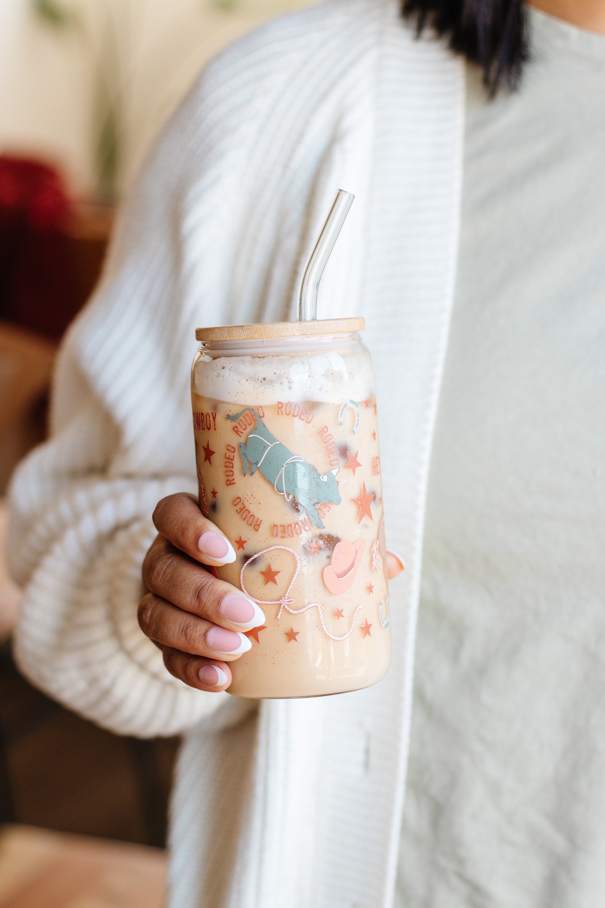 A person holding a rodeo cowboy-themed can glass with a bamboo lid and a glass straw, wearing a white sweater.