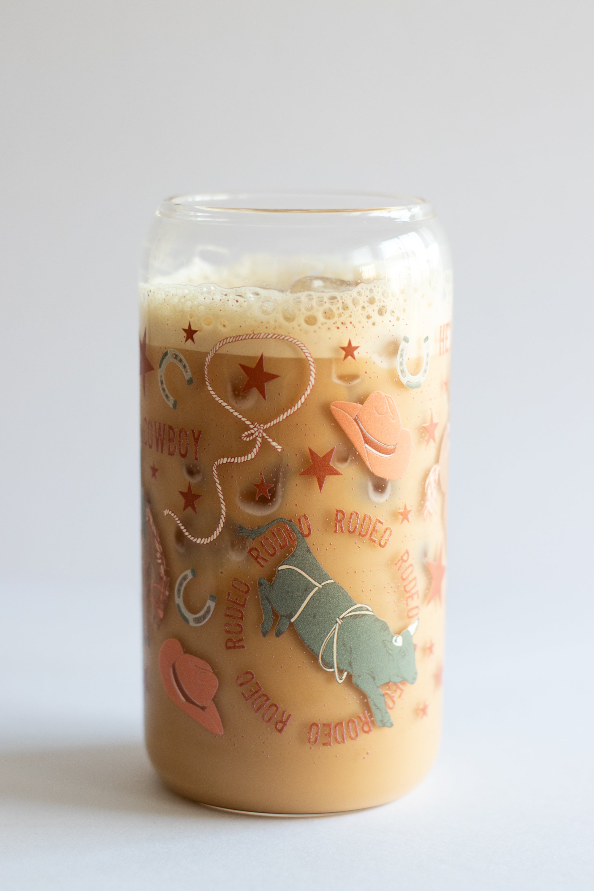 A can glass tumbler filled with iced coffee with cowboy-themed illustrations on a white background