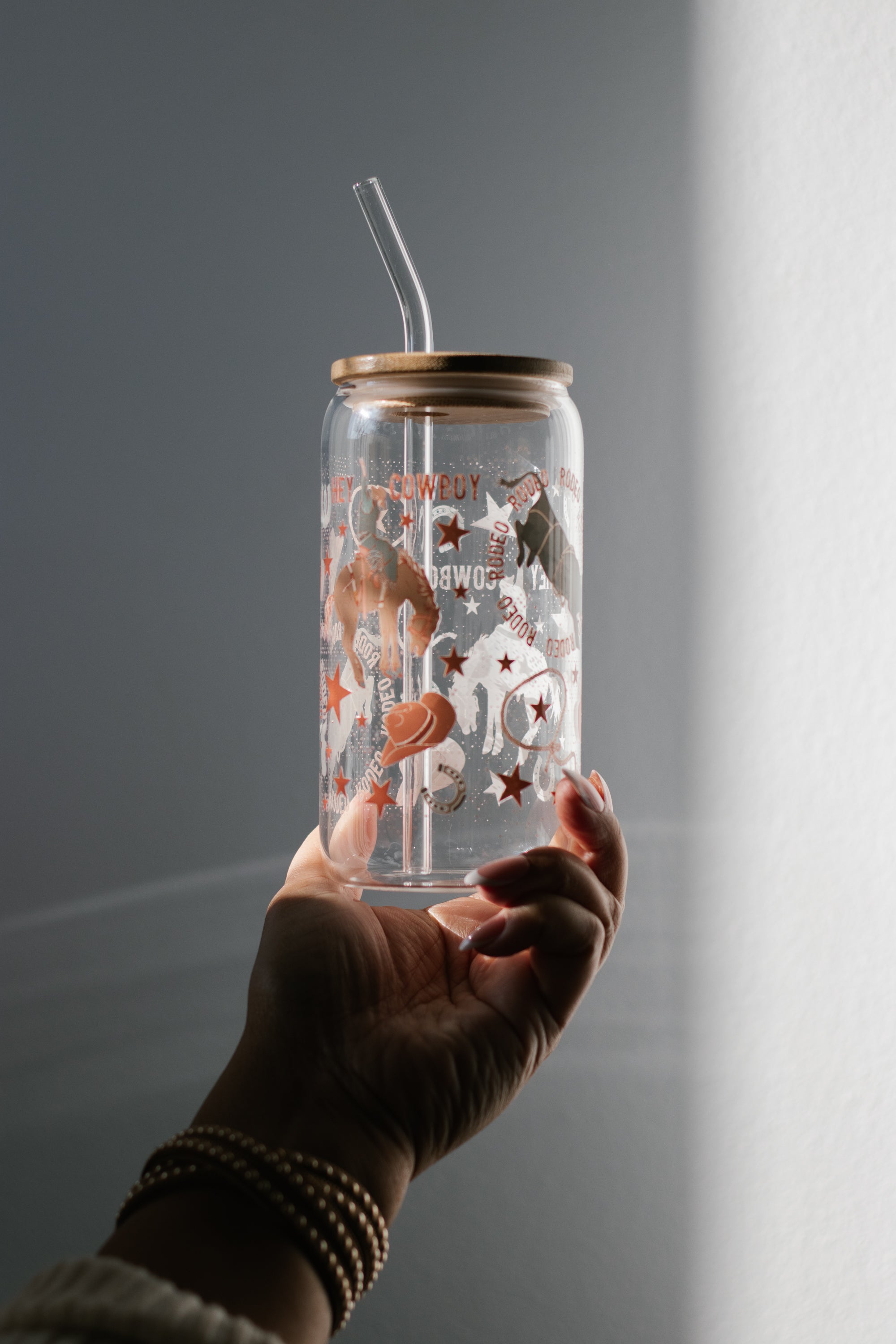 Hand holding a can glass tumbler with a bmaboo lid and a glass straw against a gray background