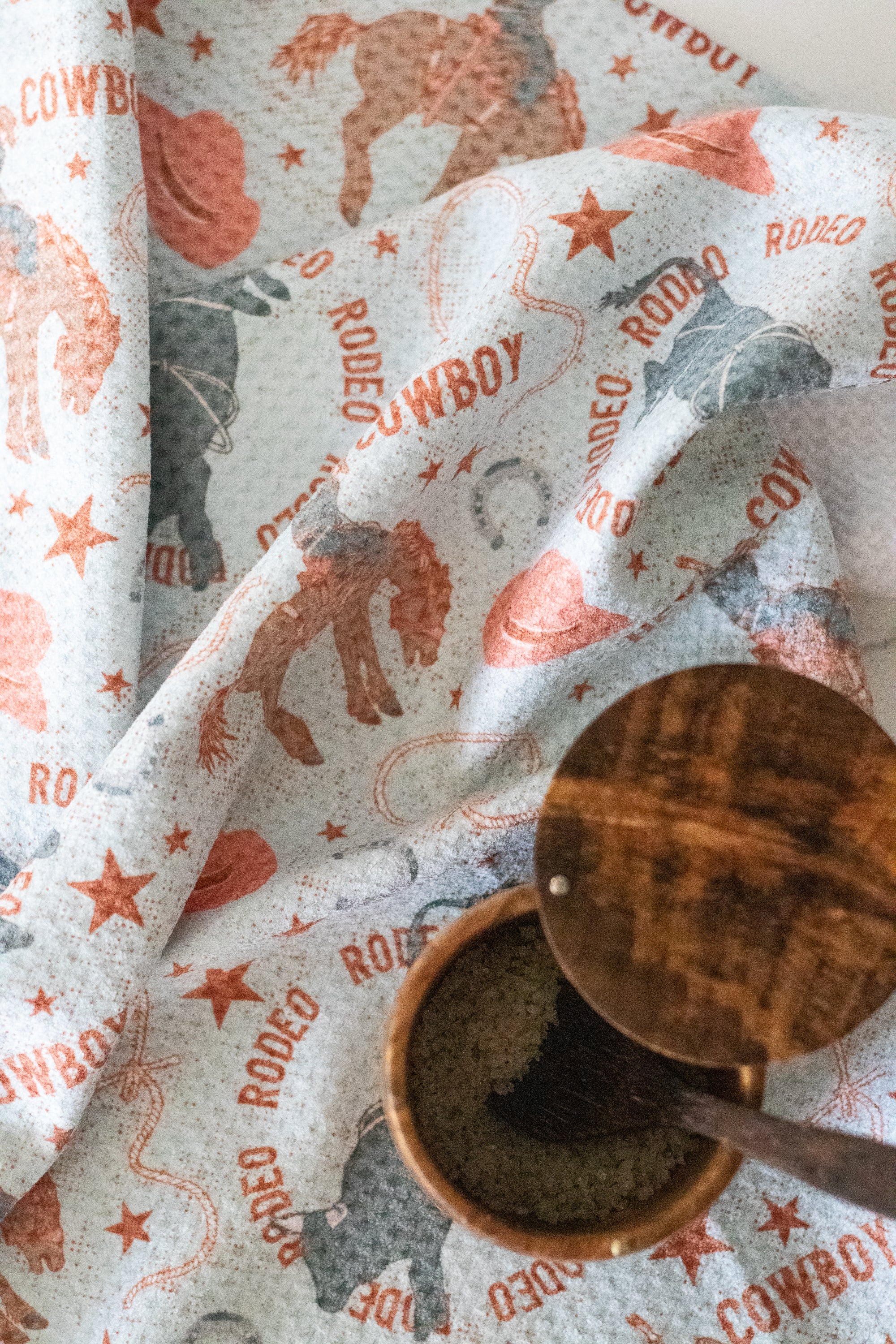 A waffle-weave kitchen tea towel with a cowboy and rodeo design on a light blue background, with a wooden salt jar and spoon, on a white surface