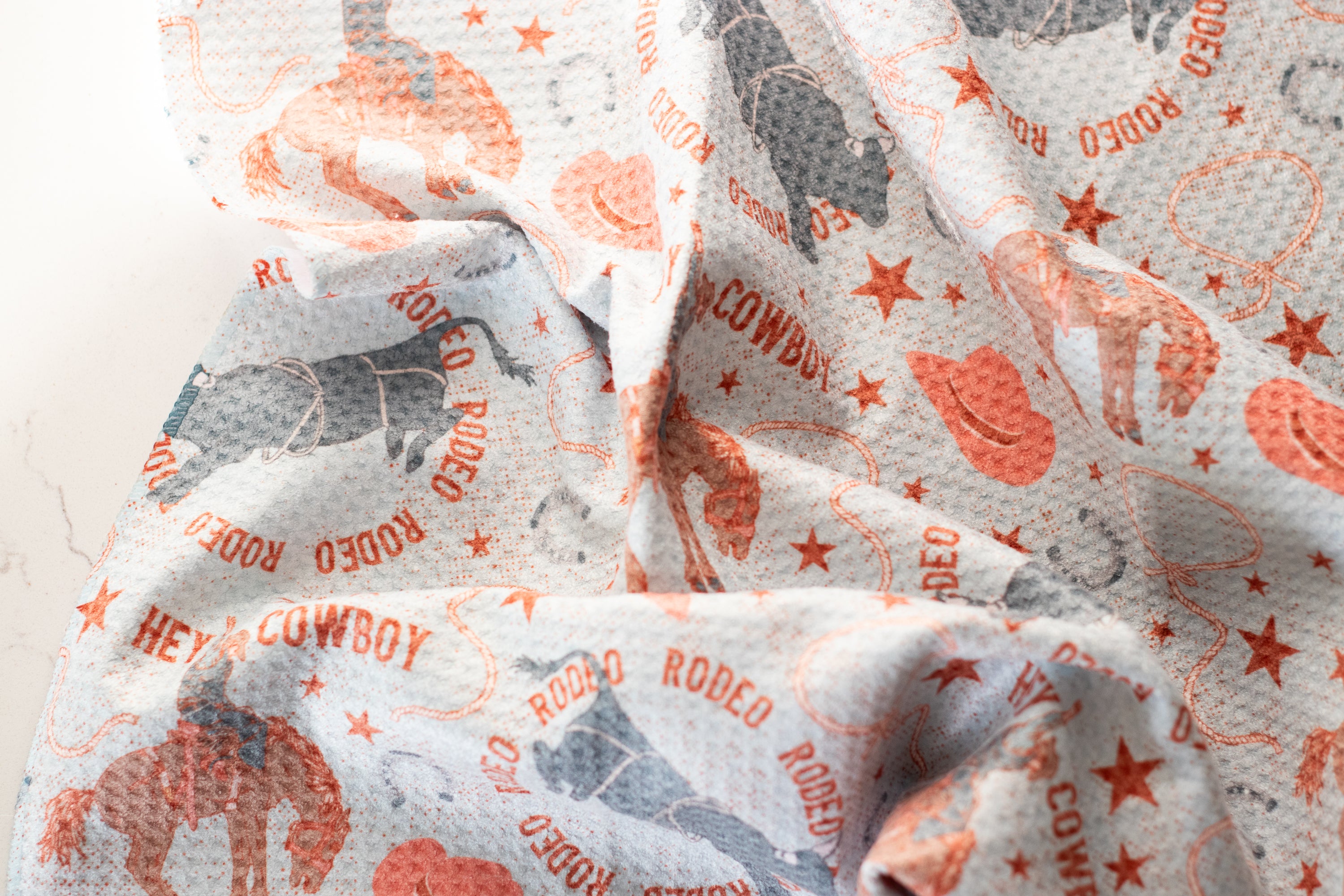 A close-up of a waffle-weave kitchen tea towel with a cowboy and rodeo design on a light blue background, on a white surface