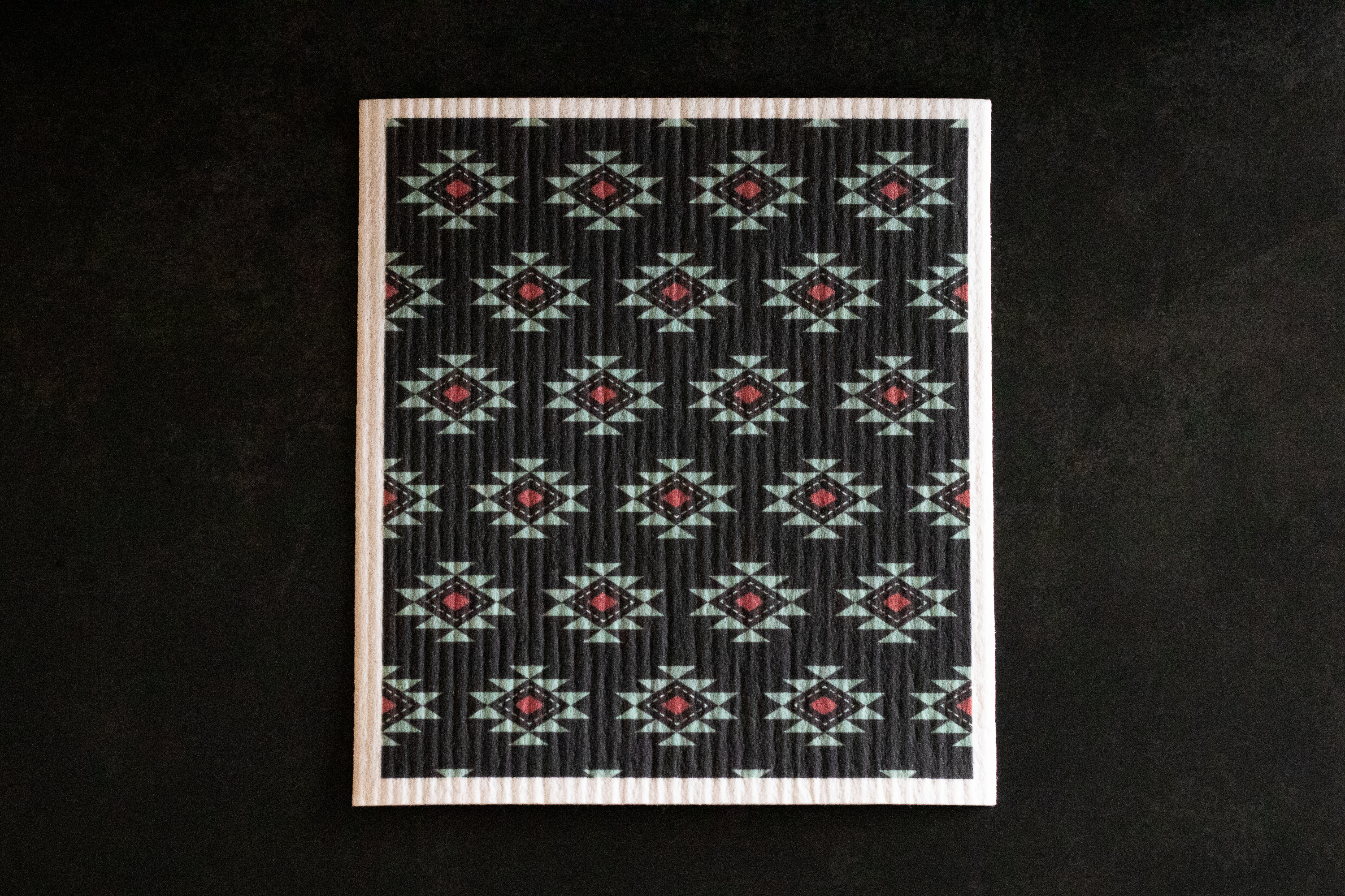 Patterned Swedish dishcloth with a Southwestern geometric design in turquoise, white, and red, on a black background.