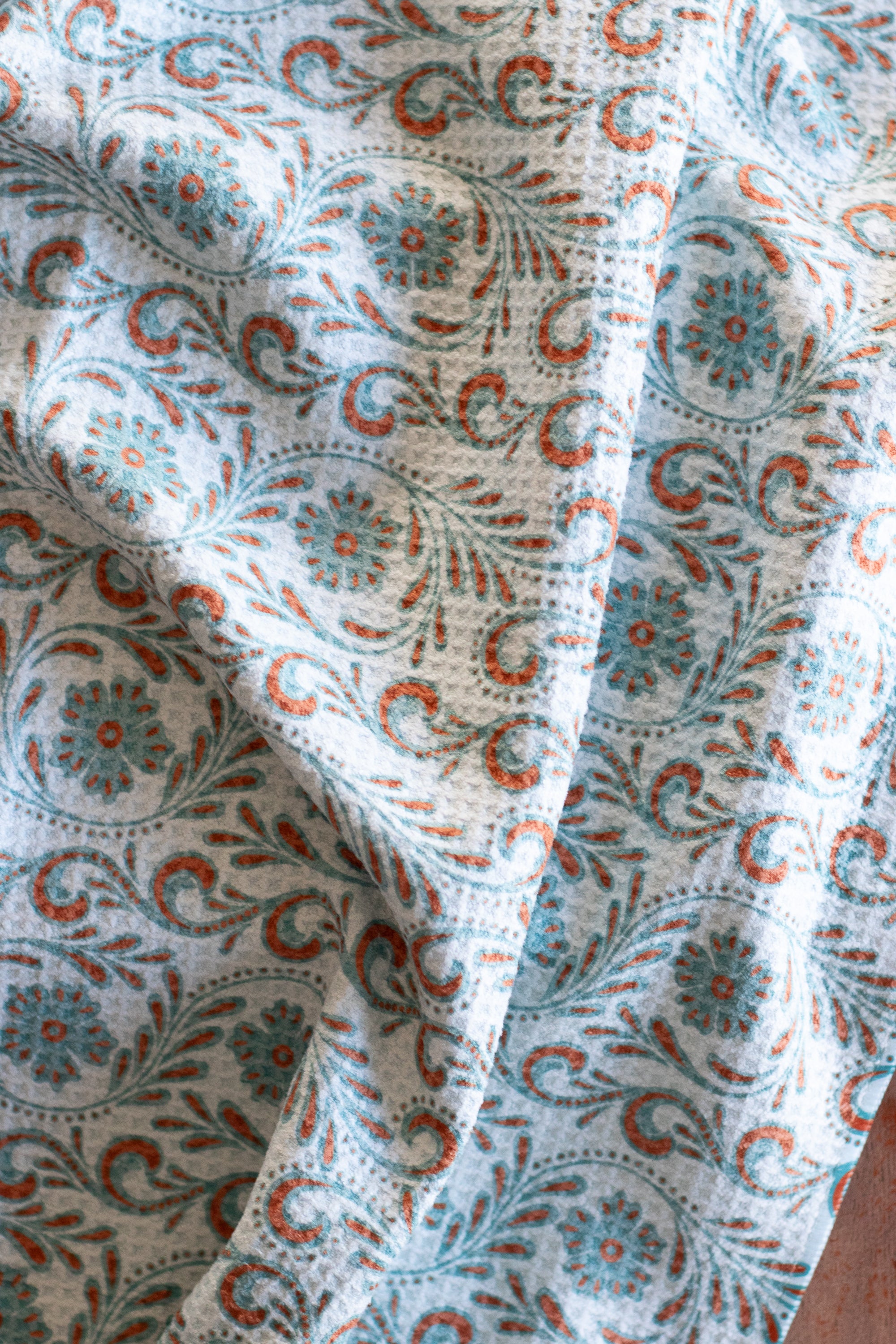 Close-up of a waffle-weave kitchen tea towel with a blue and terracotta-colored, tooled leather floral design, on a wood surface.